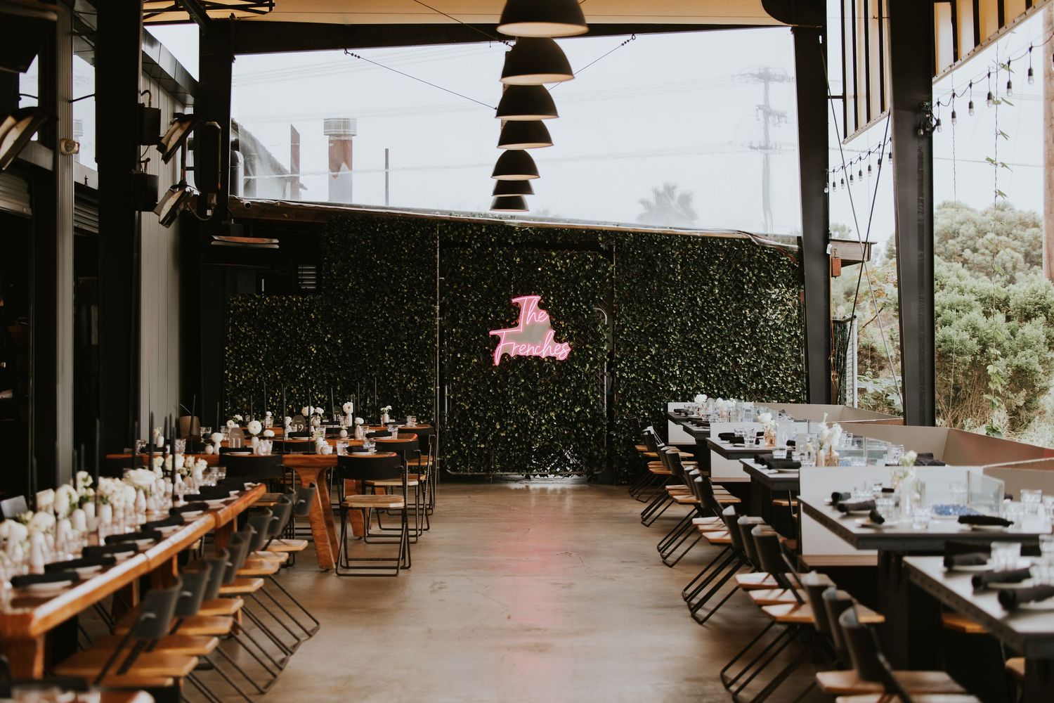 Modern restaurant interior with wooden furniture, pendant lights, and large windows overlooking greenery.