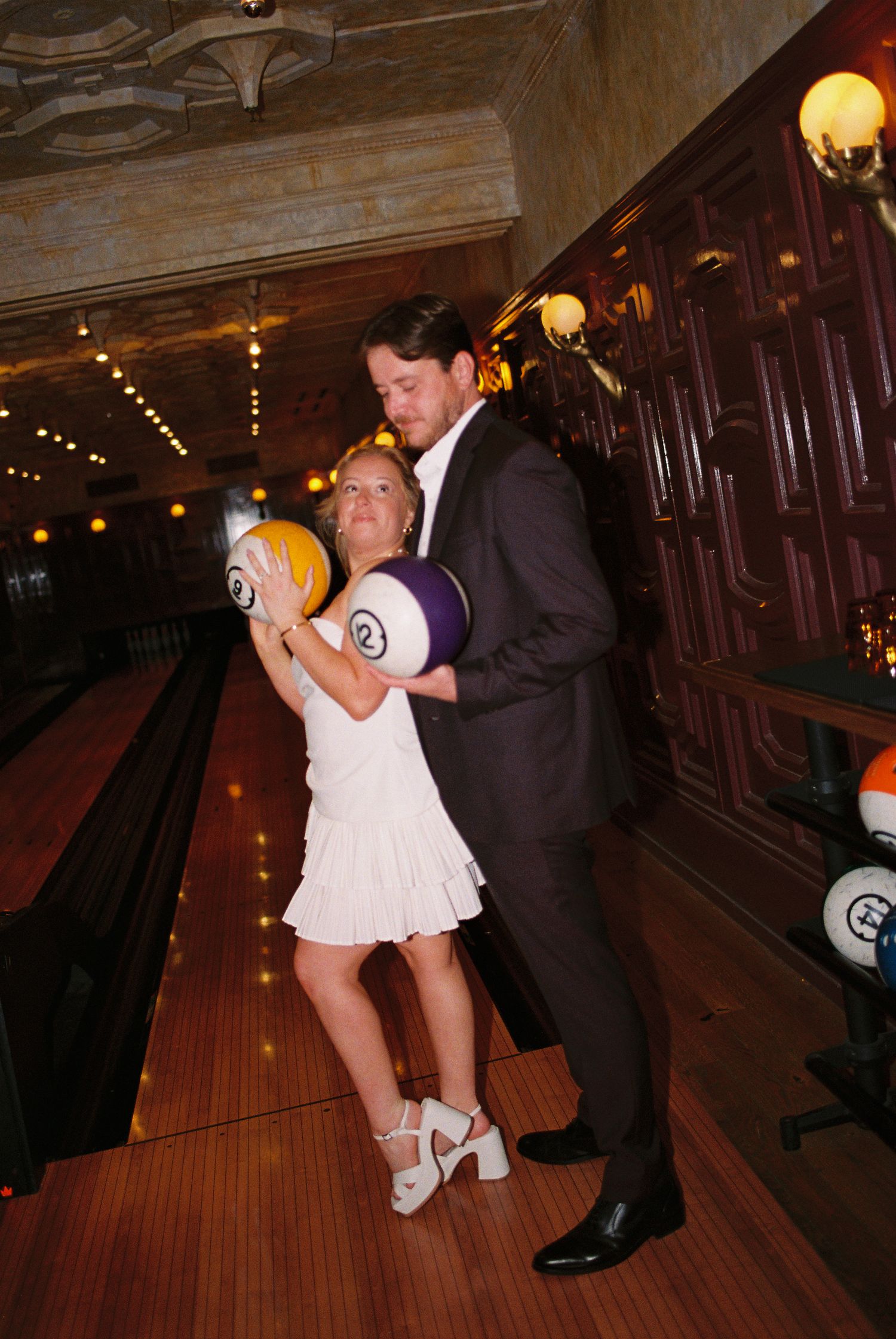 Couple in formal attire holding bowling balls at an elegant indoor bowling alley with warm lighting.