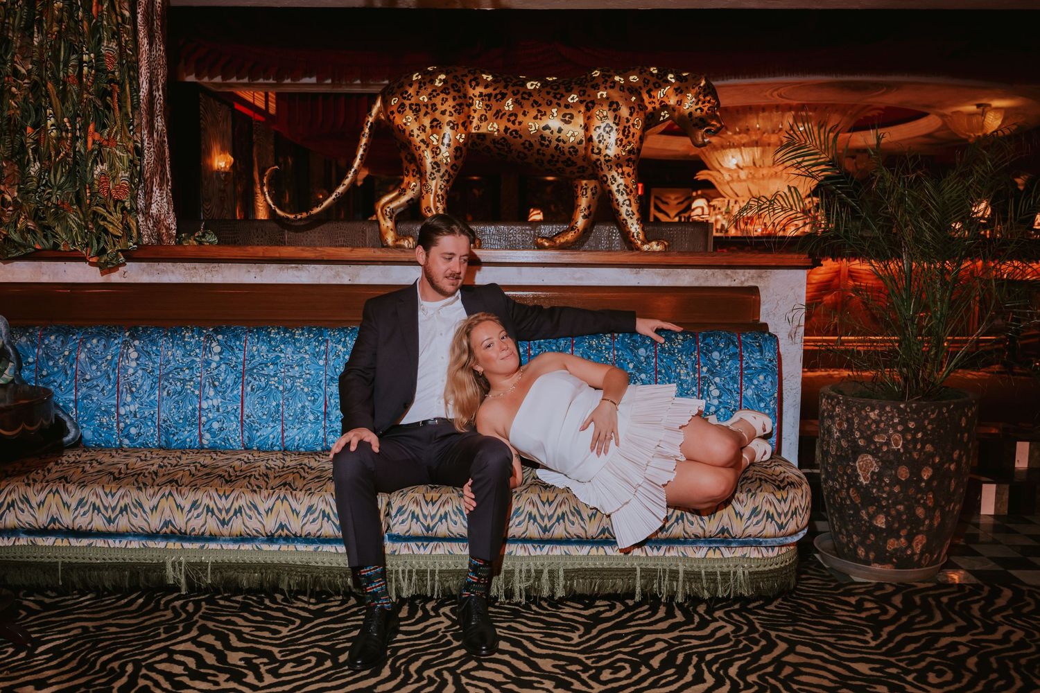 Couple in wedding attire relaxing on a patterned sofa with a gold leopard statue in a moody venue.