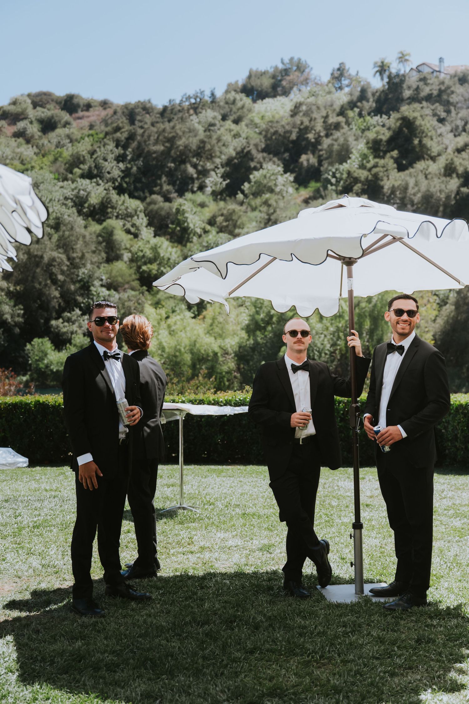 Four groomsmen in black tuxedos and sunglasses stand outdoors under a white scalloped umbrella on a sunny day.
