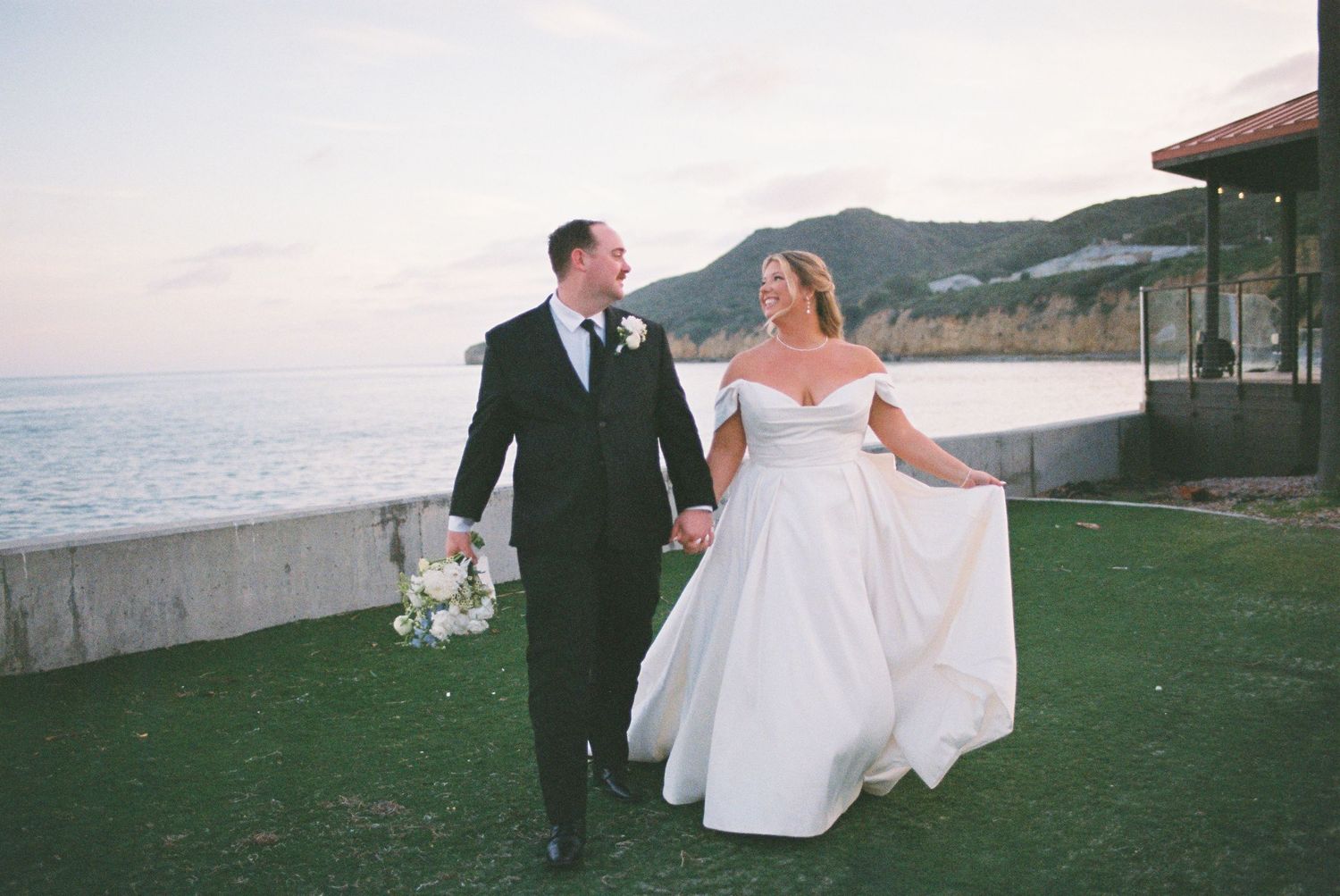 Bride and groom walk hand-in-hand on a seaside lawn at sunset, with ocean and mountains in the background.