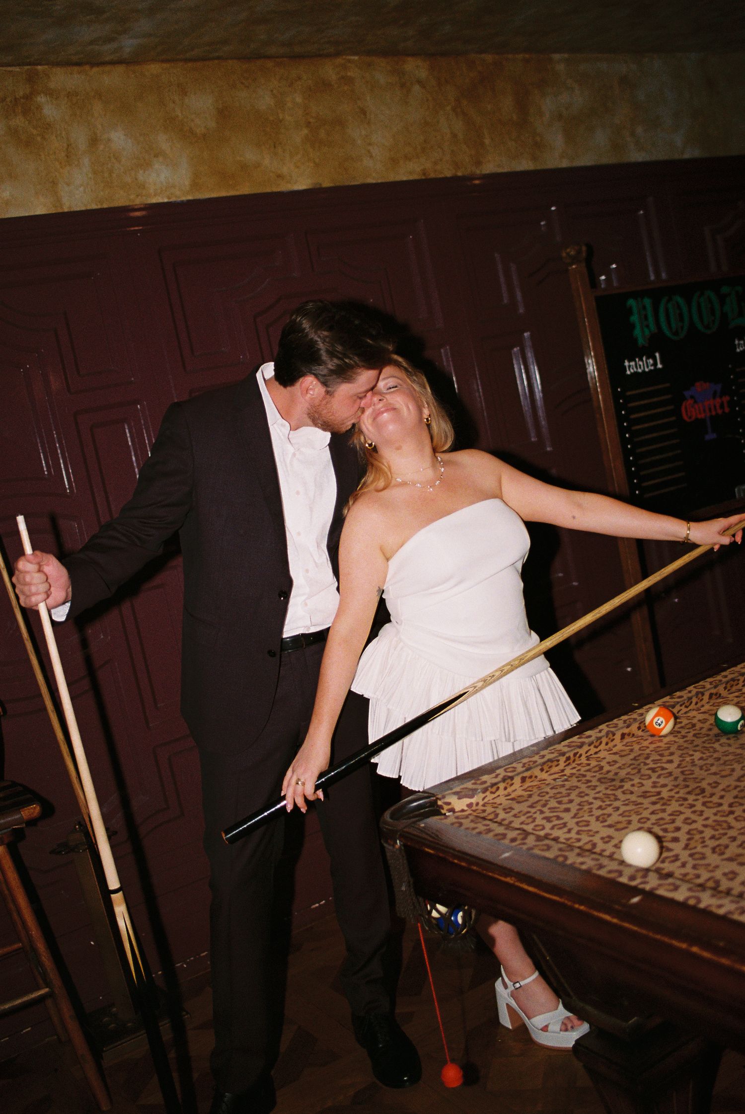 Couple sharing a kiss while playing pool, bride in white strapless dress holding cue stick at billiards table.