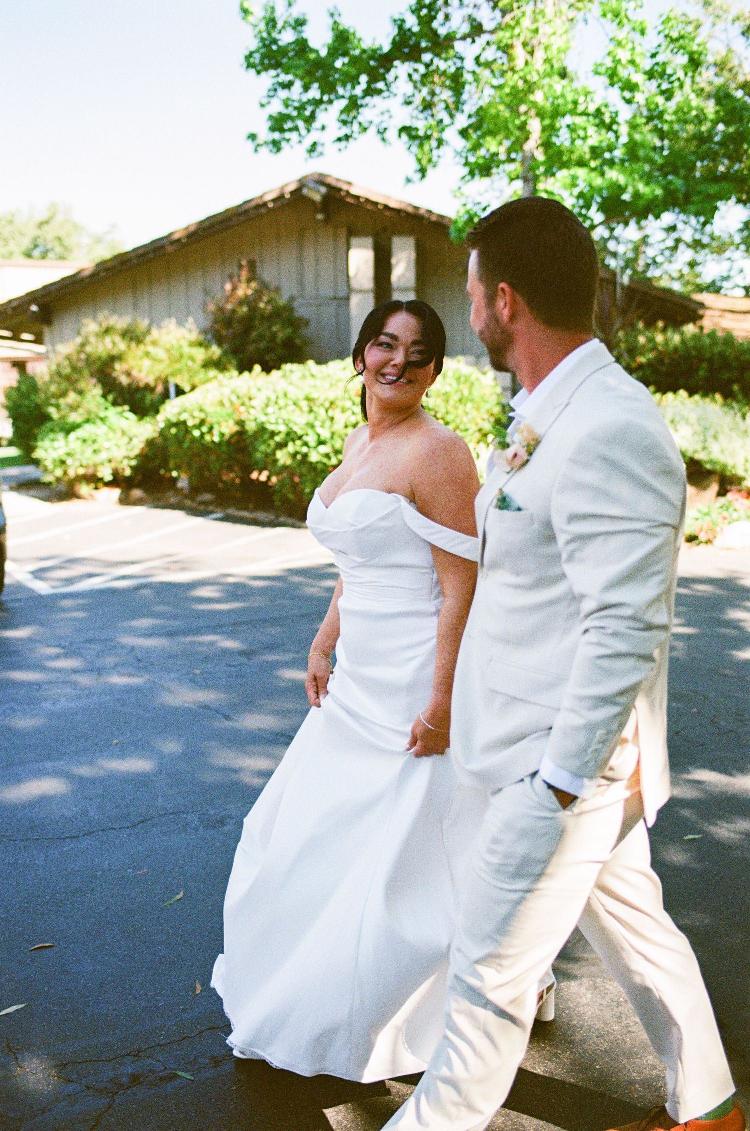 Bride in off-shoulder white gown and groom in white suit walk and laugh together on sunny wedding day.