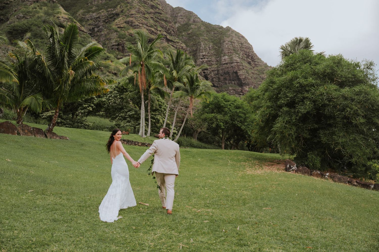 Couple walks hand in hand across green lawn with mountain landscape behind them.