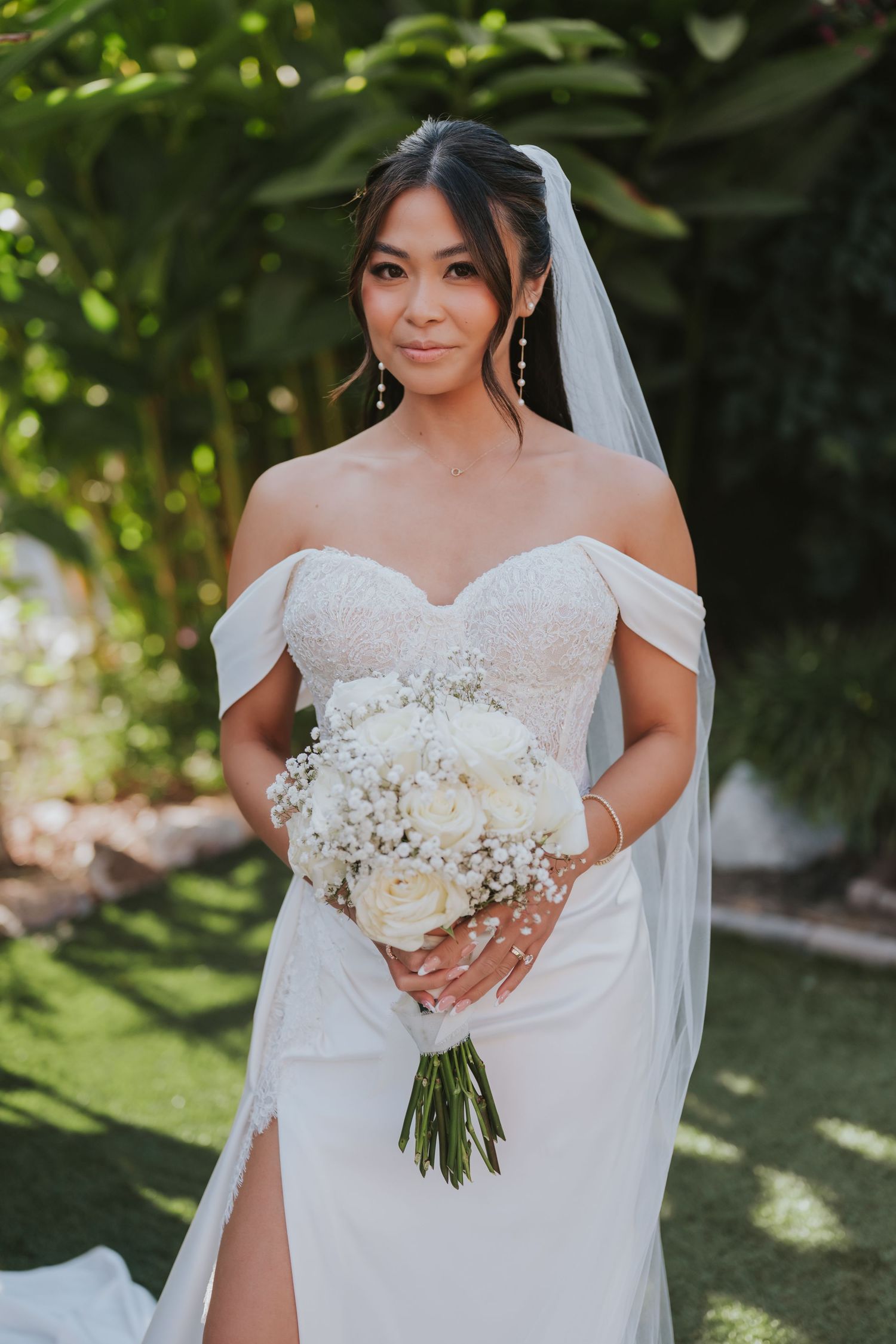 A bride in a white wedding dress holds a classic white and green bouquet in an outdoor garden setting.