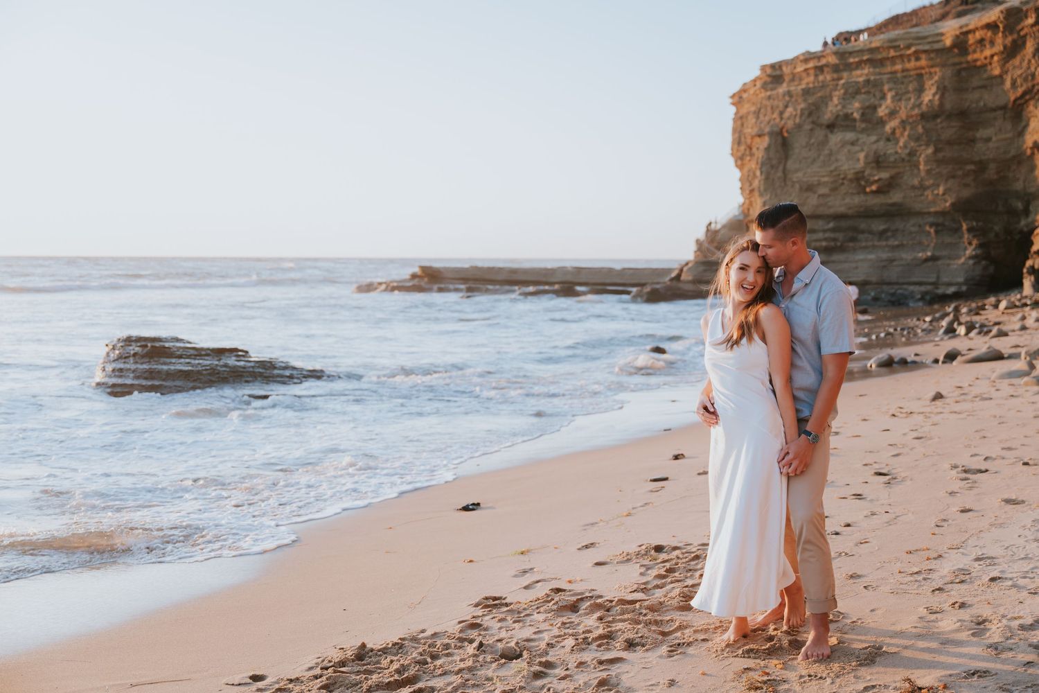 Couple embracing on a sandy beach at sunset with ocean waves and rocky cliffs in the background.