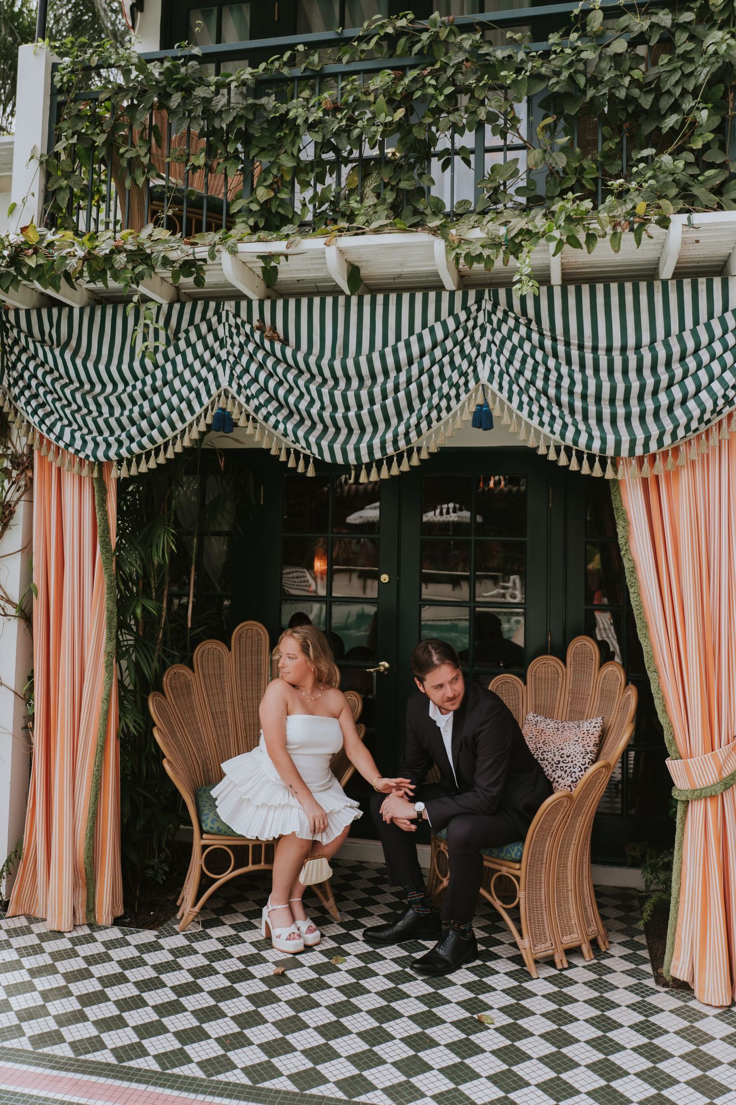 Couple in wedding attire sitting on wicker chairs under striped awning with pink curtains on checkered floor.