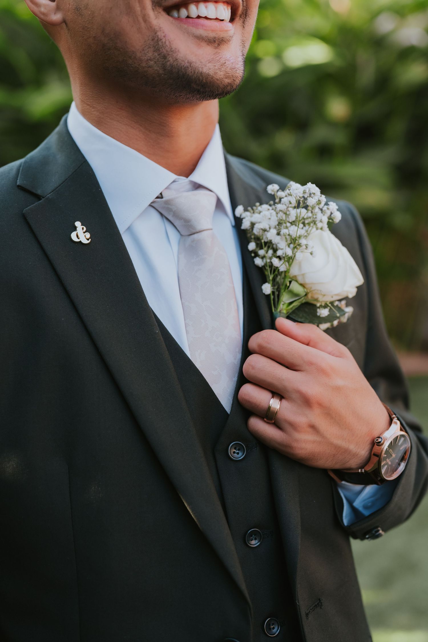Close-up view of a groom in a dark suit with white baby's breath boutonniere against a green garden background.