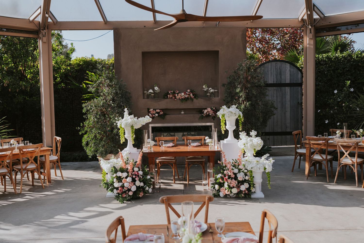Elegant outdoor wedding reception setup with wooden furniture and floral arrangements under a covered patio.