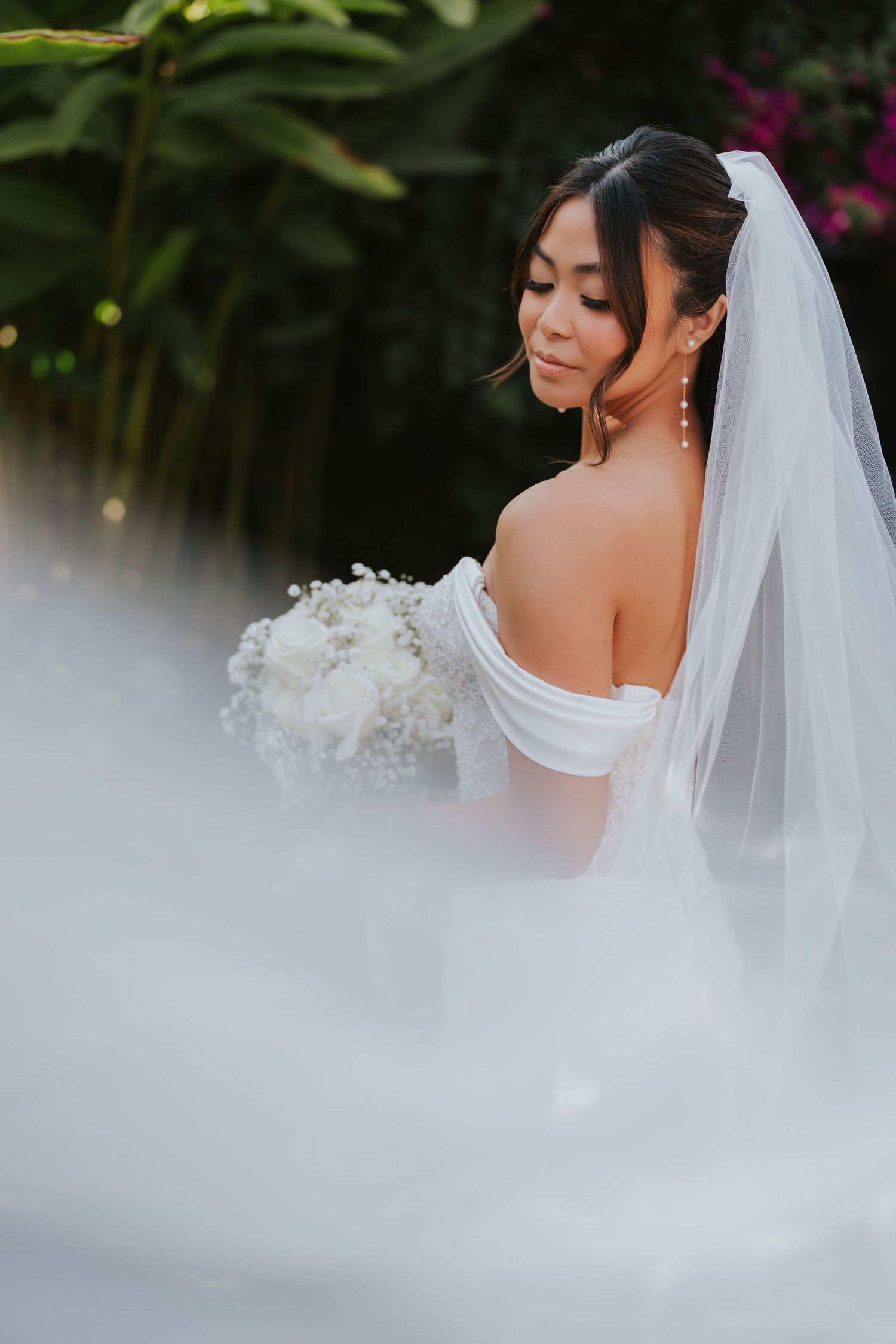 A bride in an off-shoulder wedding dress holds a white bouquet while wearing a flowing veil against a blurred background.