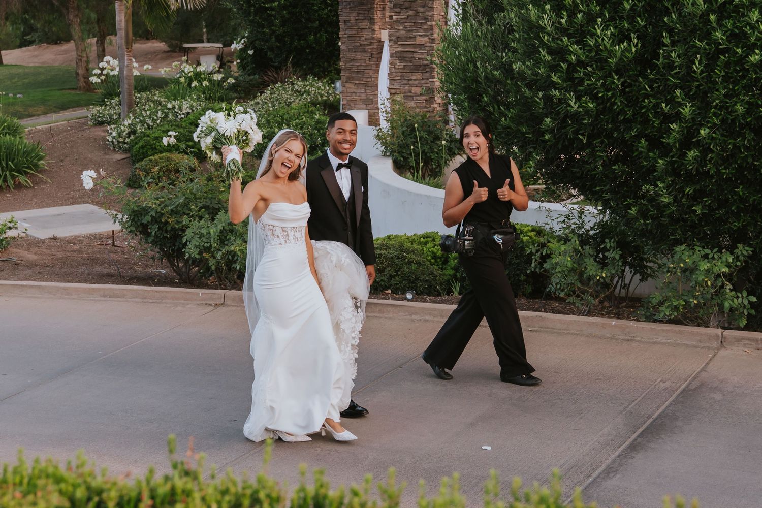 Bride and groom walking outdoors at wedding venue with bridesmaid holding dress train, lush greenery background.