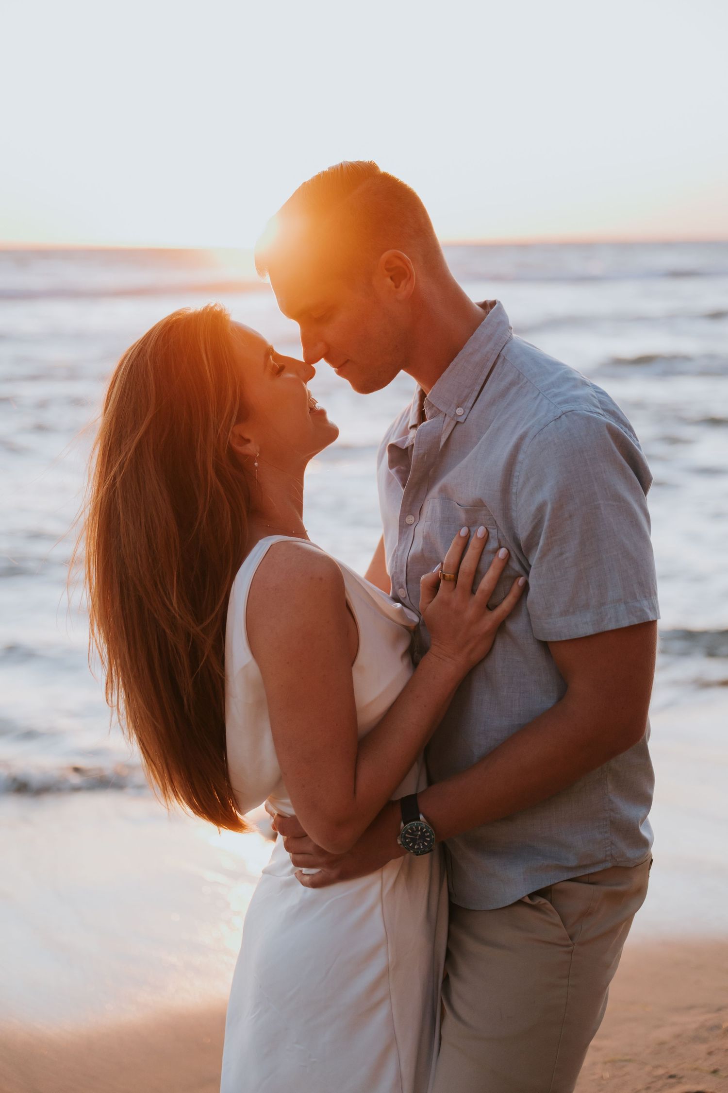 Couple embracing on a beach at golden sunset, woman in white dress and man in gray shirt sharing a romantic moment.