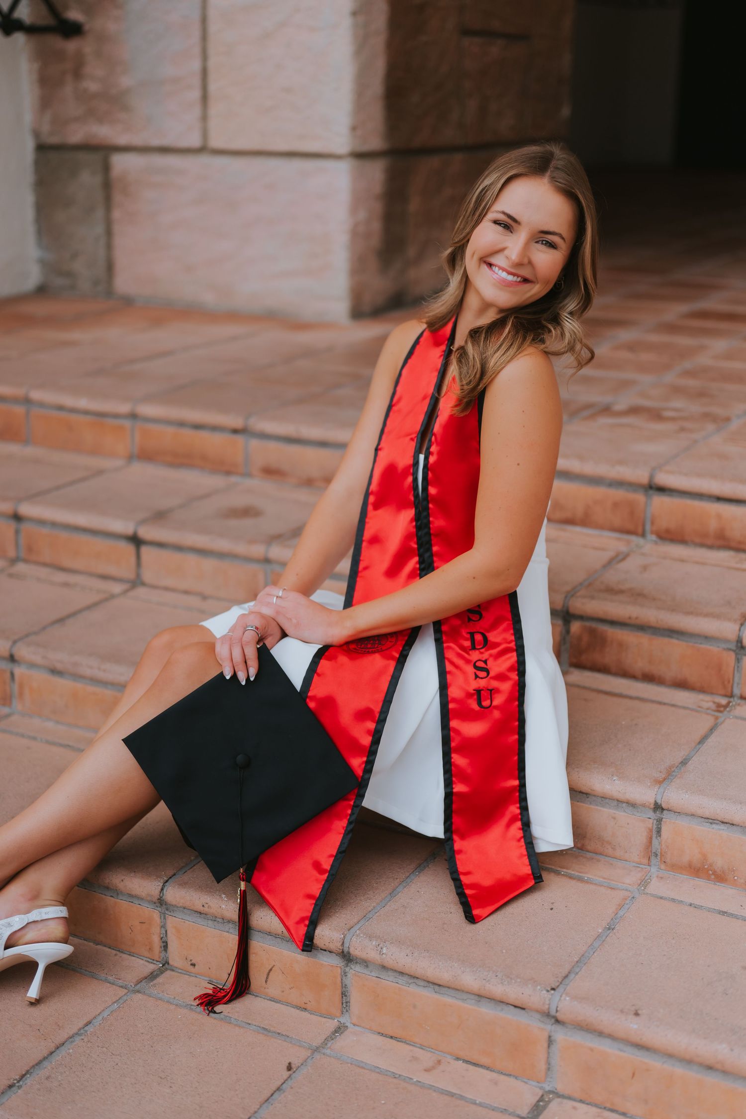 Smiling female graduate in red SDSU sash sits on brick steps holding black mortarboard cap.