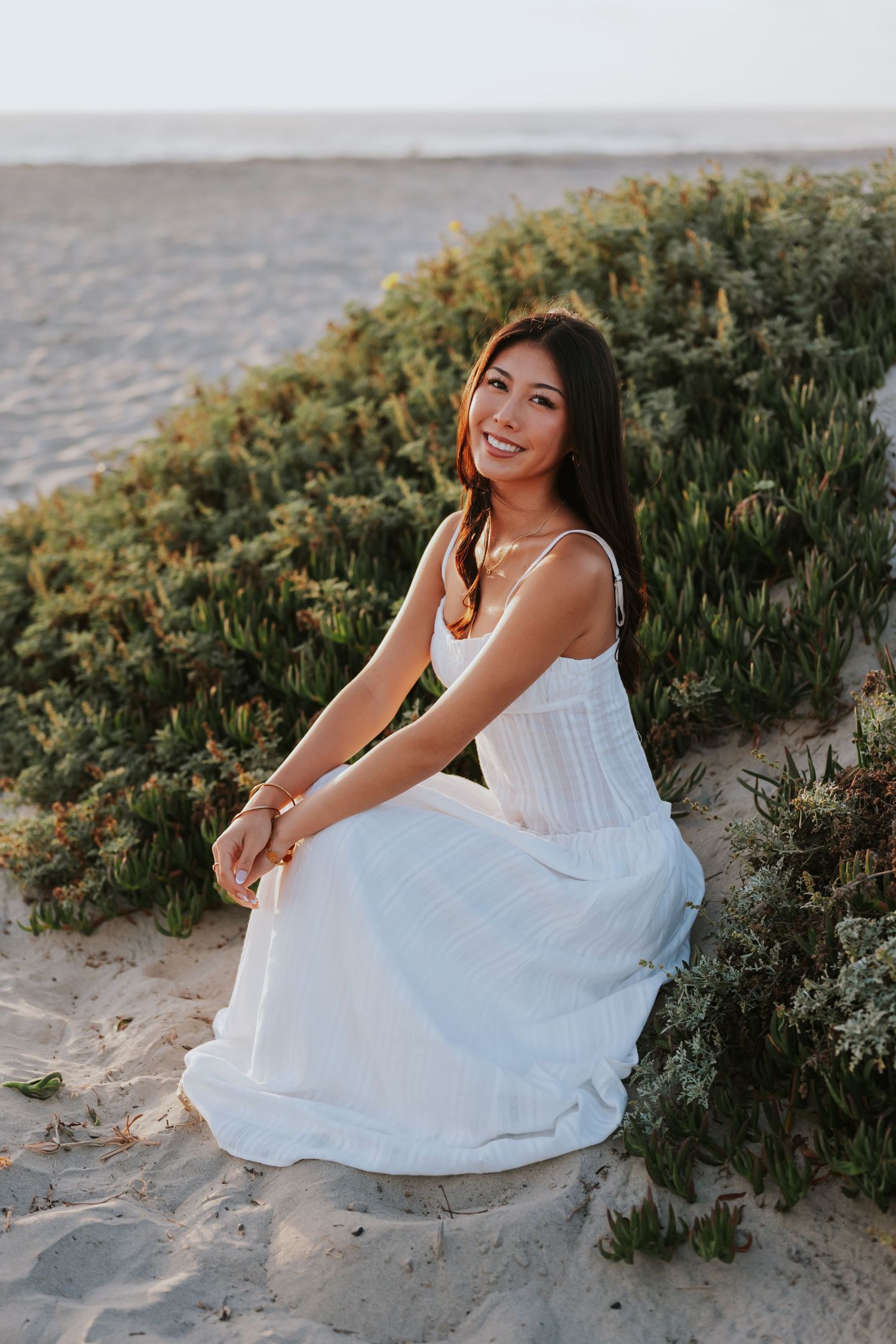 Young woman in white sundress sitting on sandy beach dunes, smiling warmly with green coastal shrubs behind her.