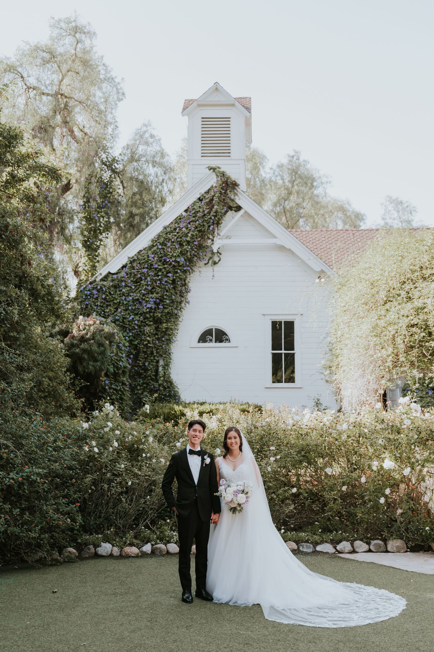 Wedding couple poses in front of charming white chapel with ivy-covered walls and garden setting.