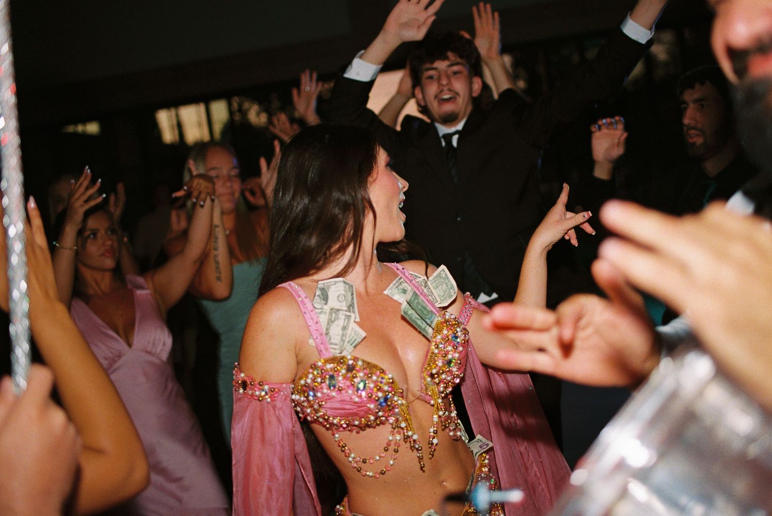 Woman in pink beaded crop top with money tucked in, dancing at a lively party with guests celebrating.