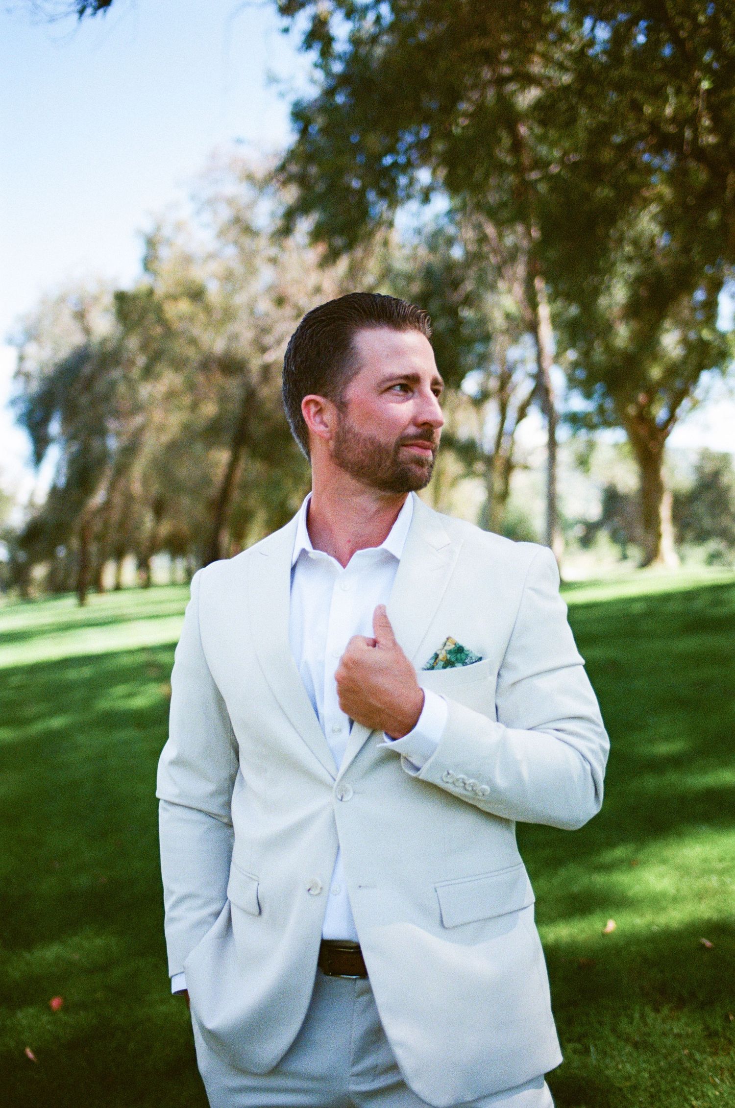 Handsome groom in cream suit with floral pocket square stands on a lush green lawn outdoors.