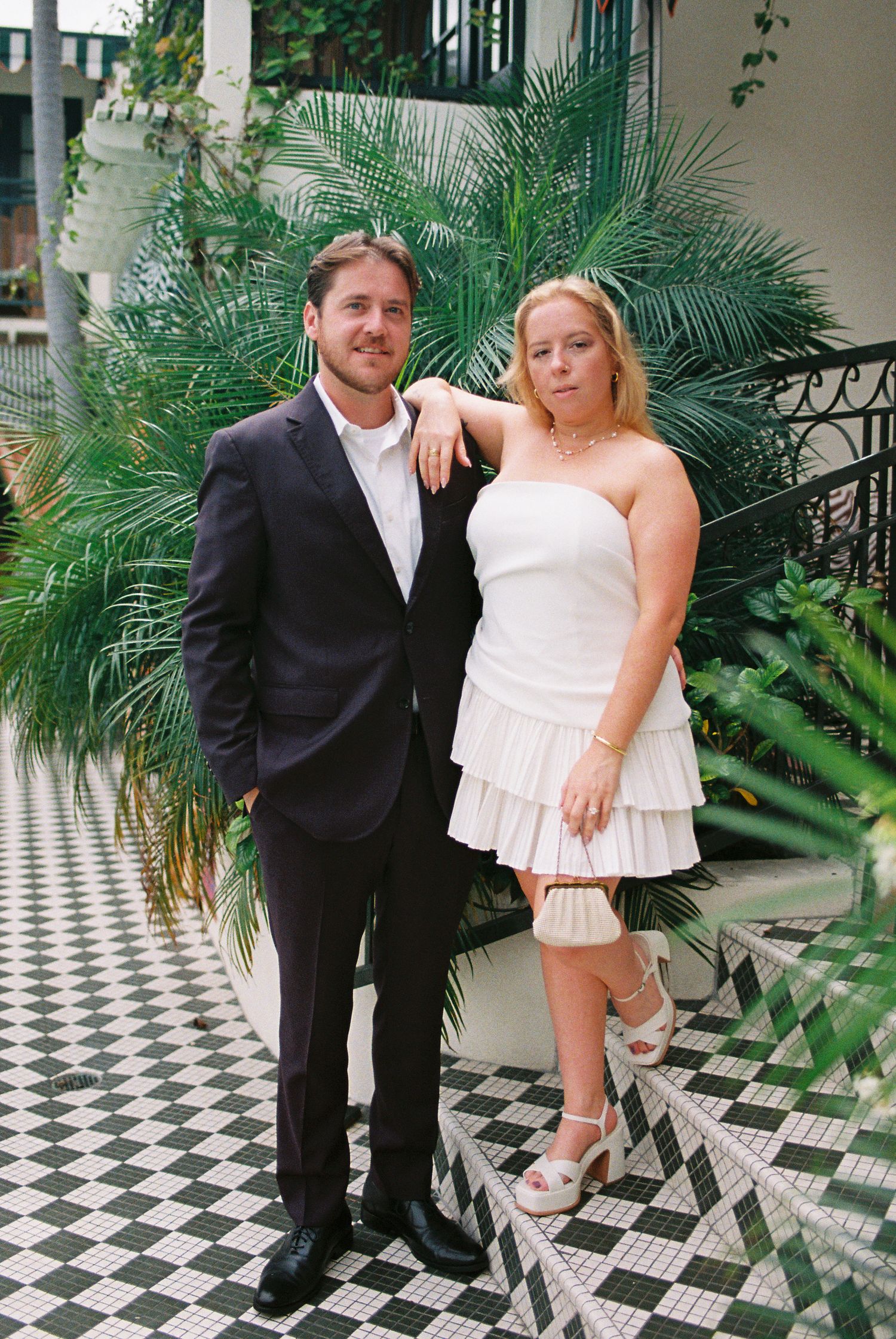 Couple posing indoors, man in dark suit, woman in white strapless mini dress, tropical plants and checkered floor.