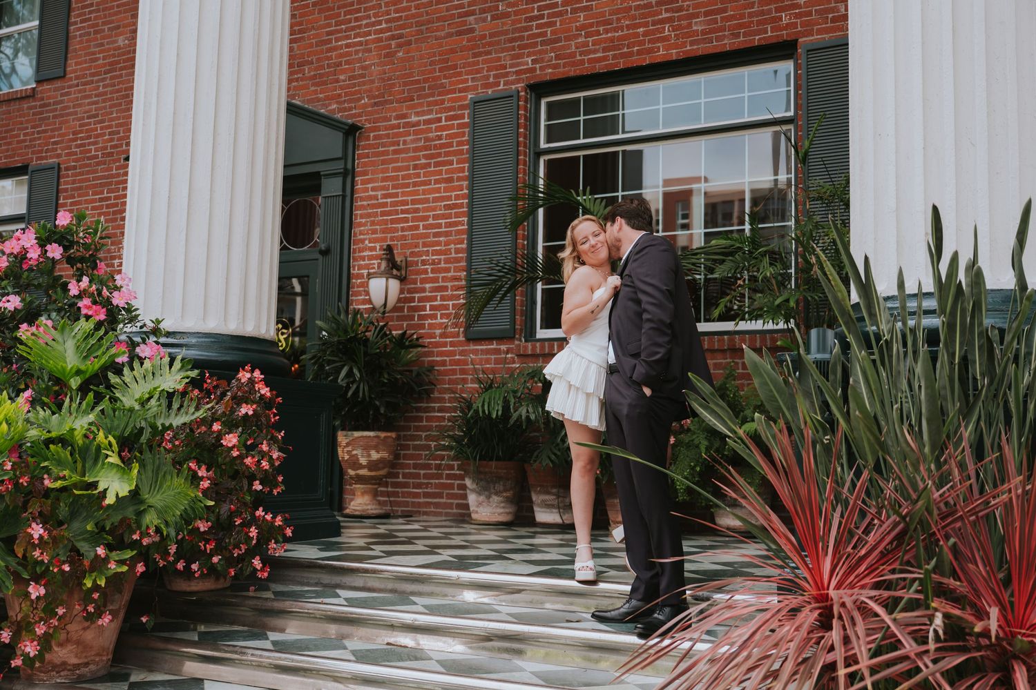 Wedding couple sharing a kiss outside a brick building entrance surrounded by lush tropical plants.