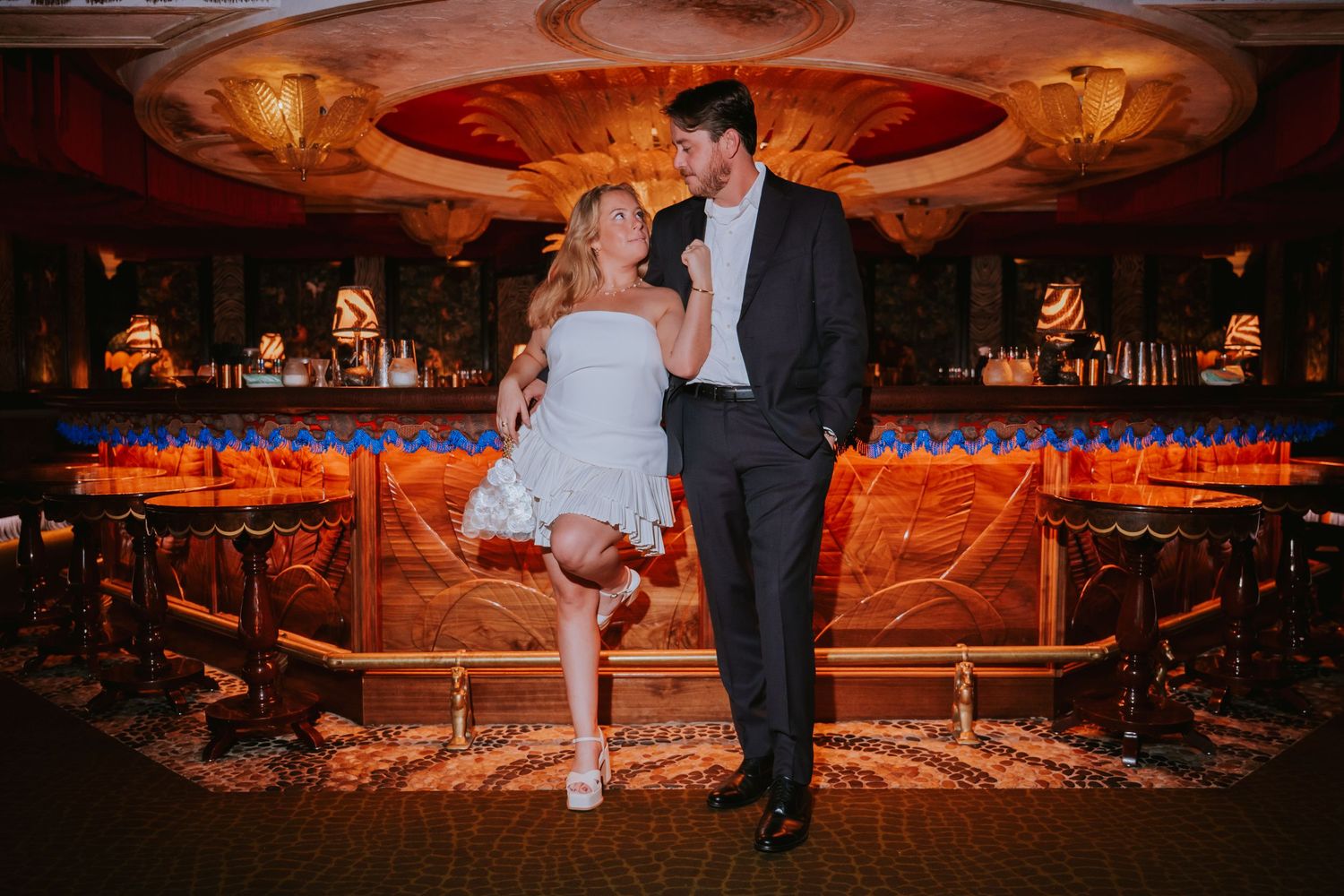 Couple posing elegantly in a luxurious bar venue, woman in white dress and man in dark suit sharing a romantic moment.