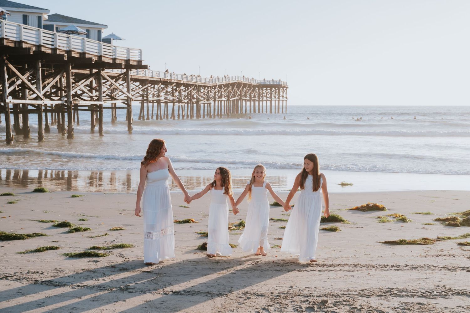 Four girls in white dresses hold hands walking on a sandy beach near a wooden pier on a sunny day.