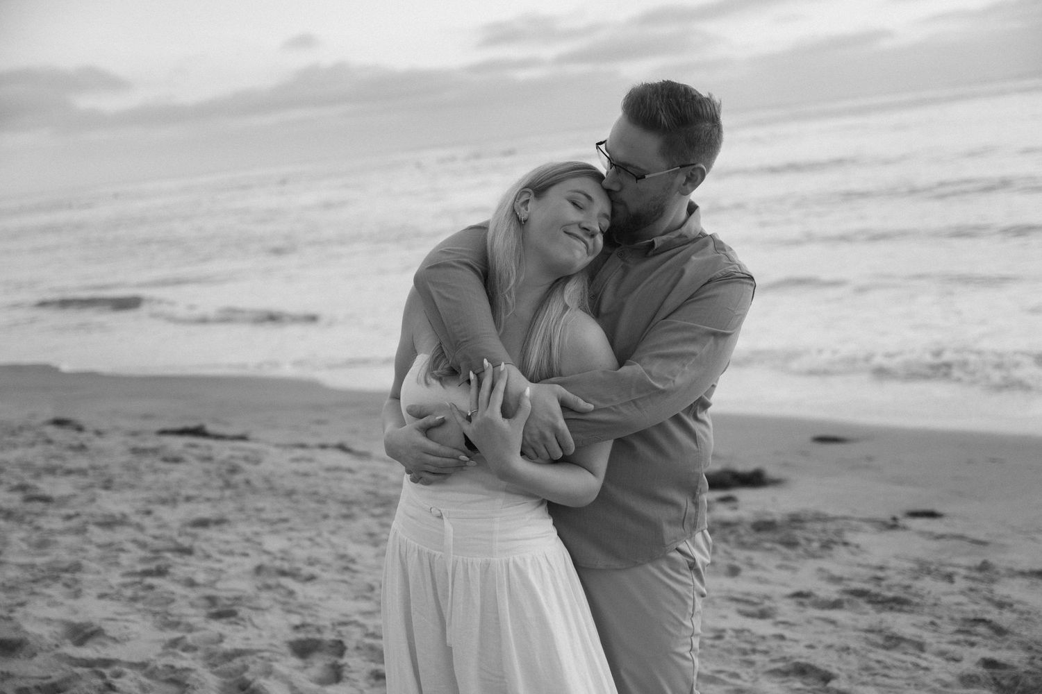 Black and white photo of a couple embracing on a beach at sunset, woman smiling with eyes closed.