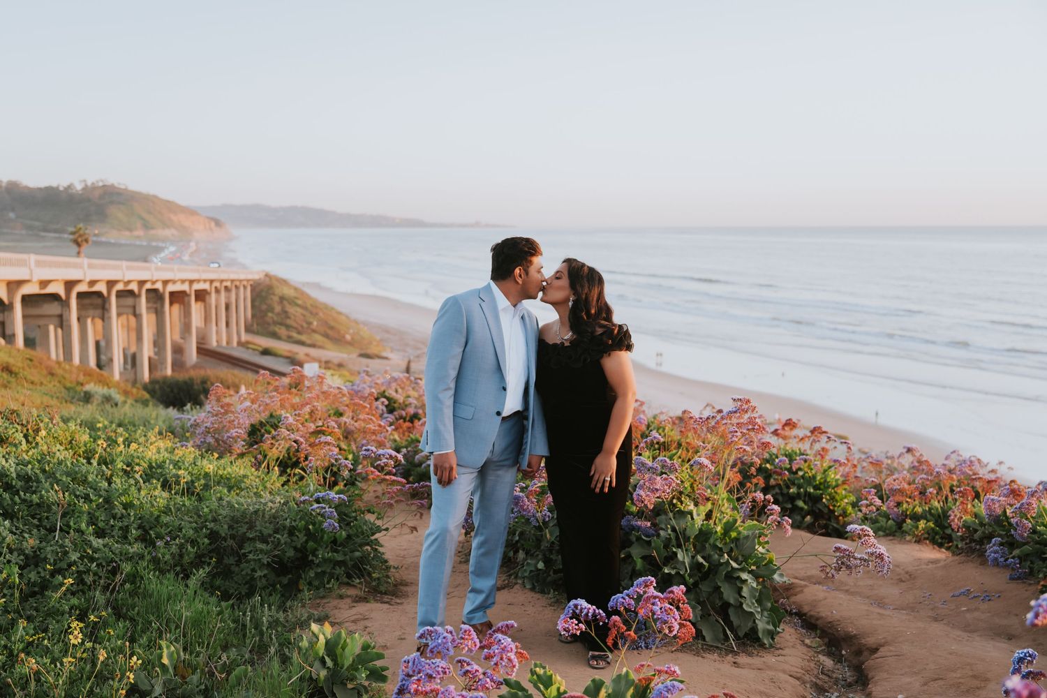 Couple kissing at sunset on coastal bluff with ocean and bridge views, surrounded by colorful flowers.