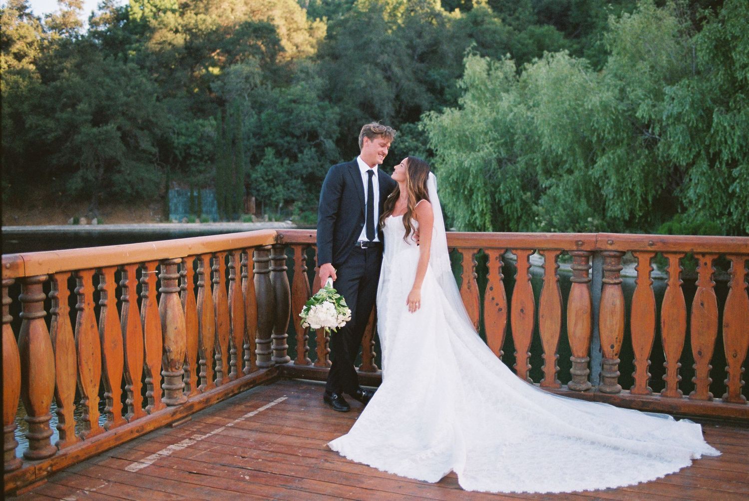 Bride and groom posing on a wooden deck surrounded by lush green trees on their wedding day.