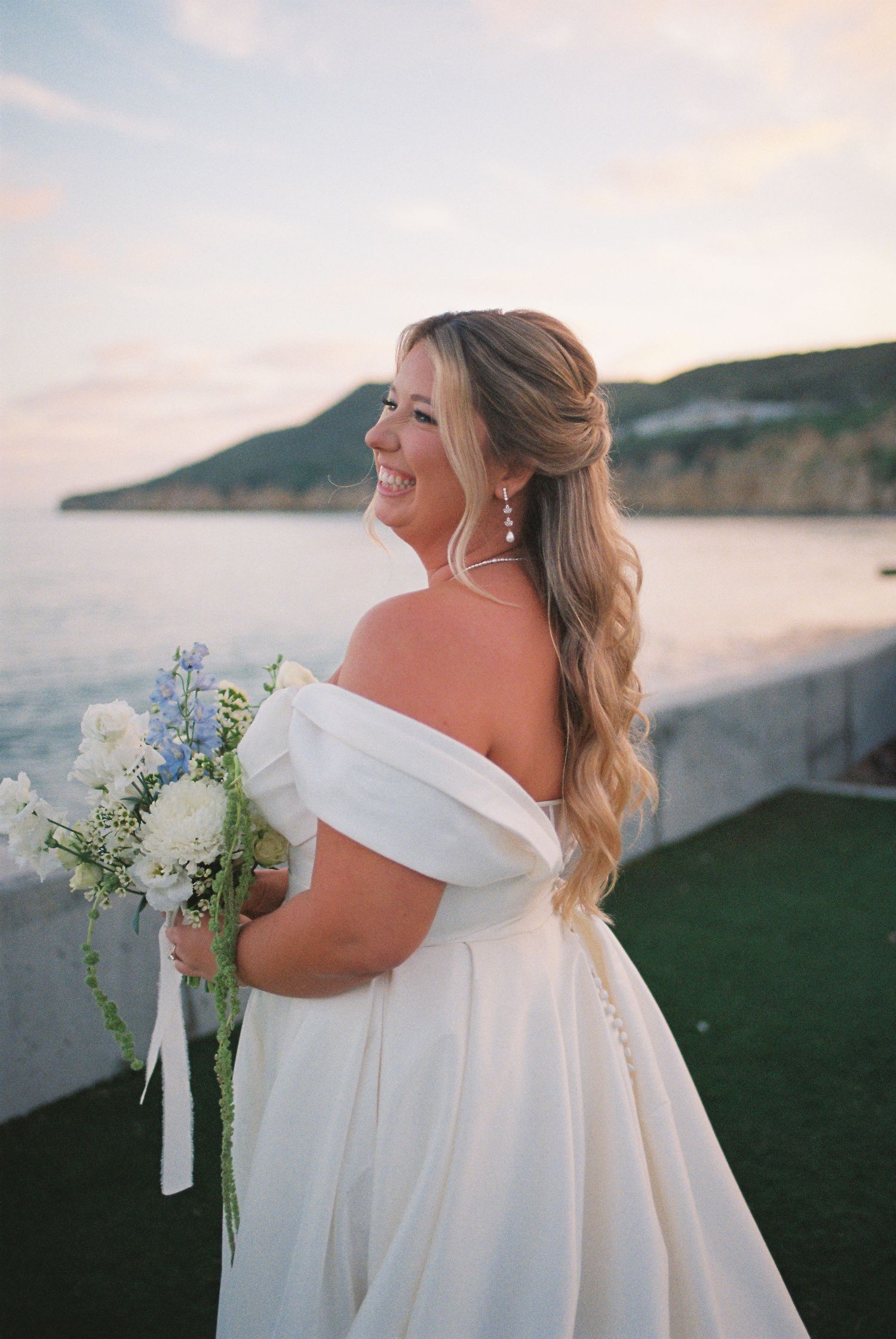 Smiling bride in off-shoulder white gown holds blue and white bouquet with ocean and hillside backdrop at sunset.