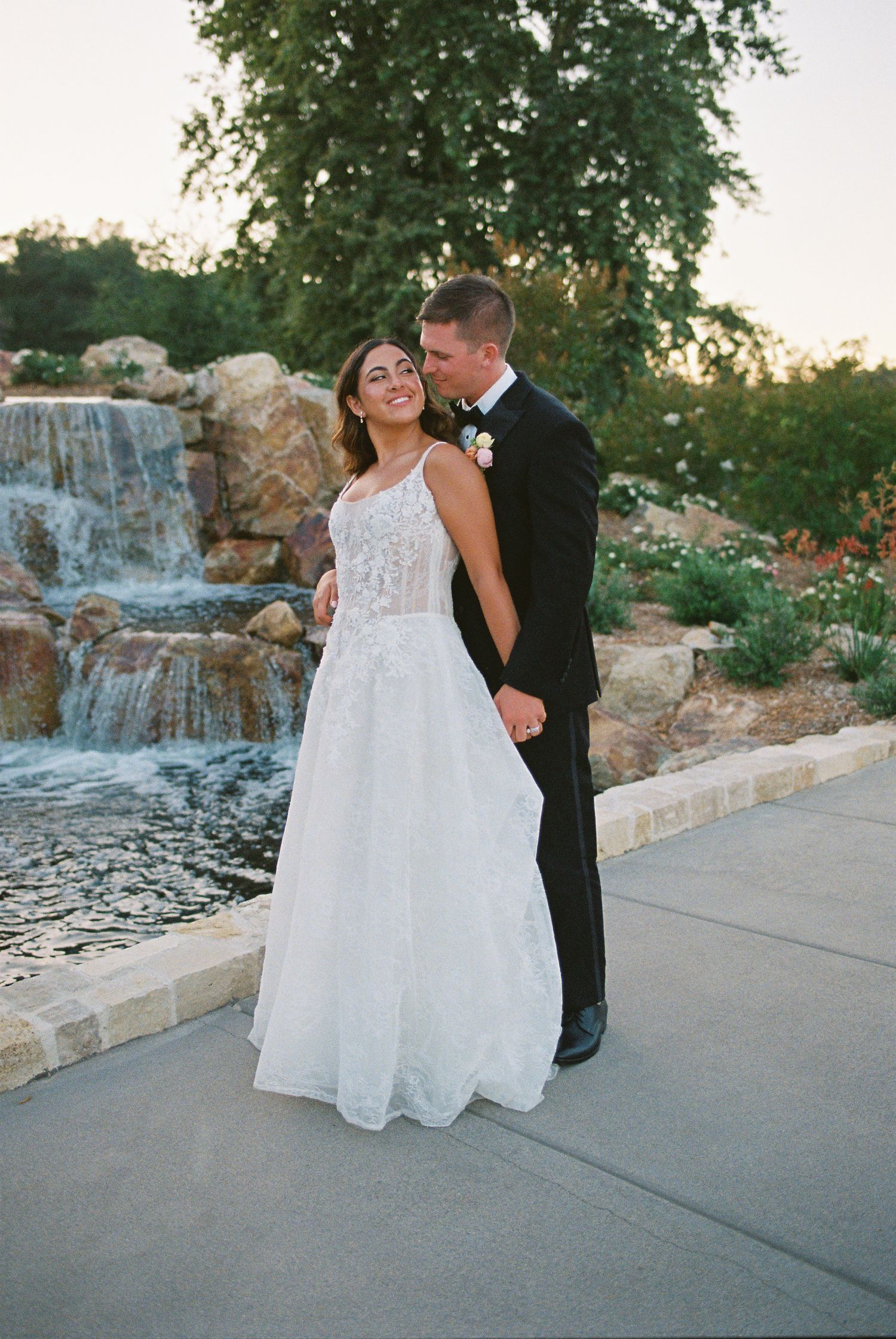 Bride in white lace gown and groom in black tuxedo embrace near a stone waterfall at golden hour.