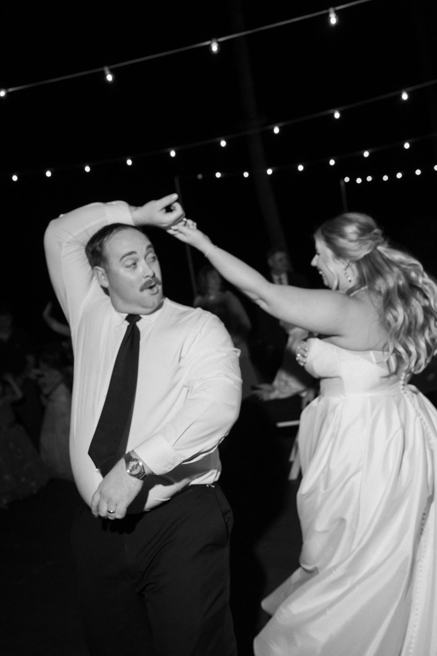 Black and white photo of a bride and groom dancing joyfully at their wedding reception under string lights.