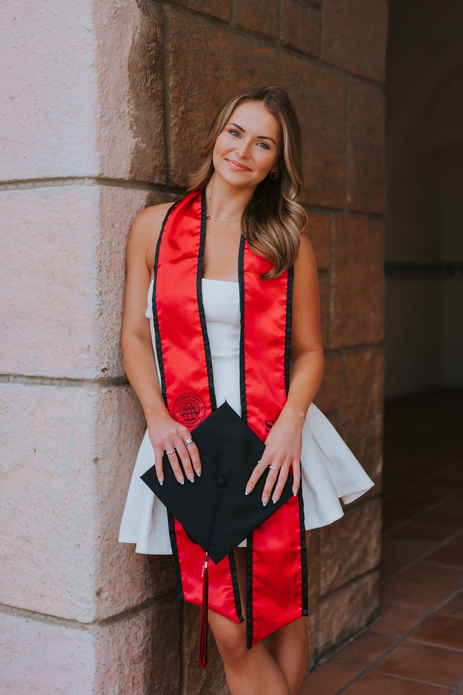 Young woman in white dress with red graduation stole holds cap, posing by stone column for grad photos.