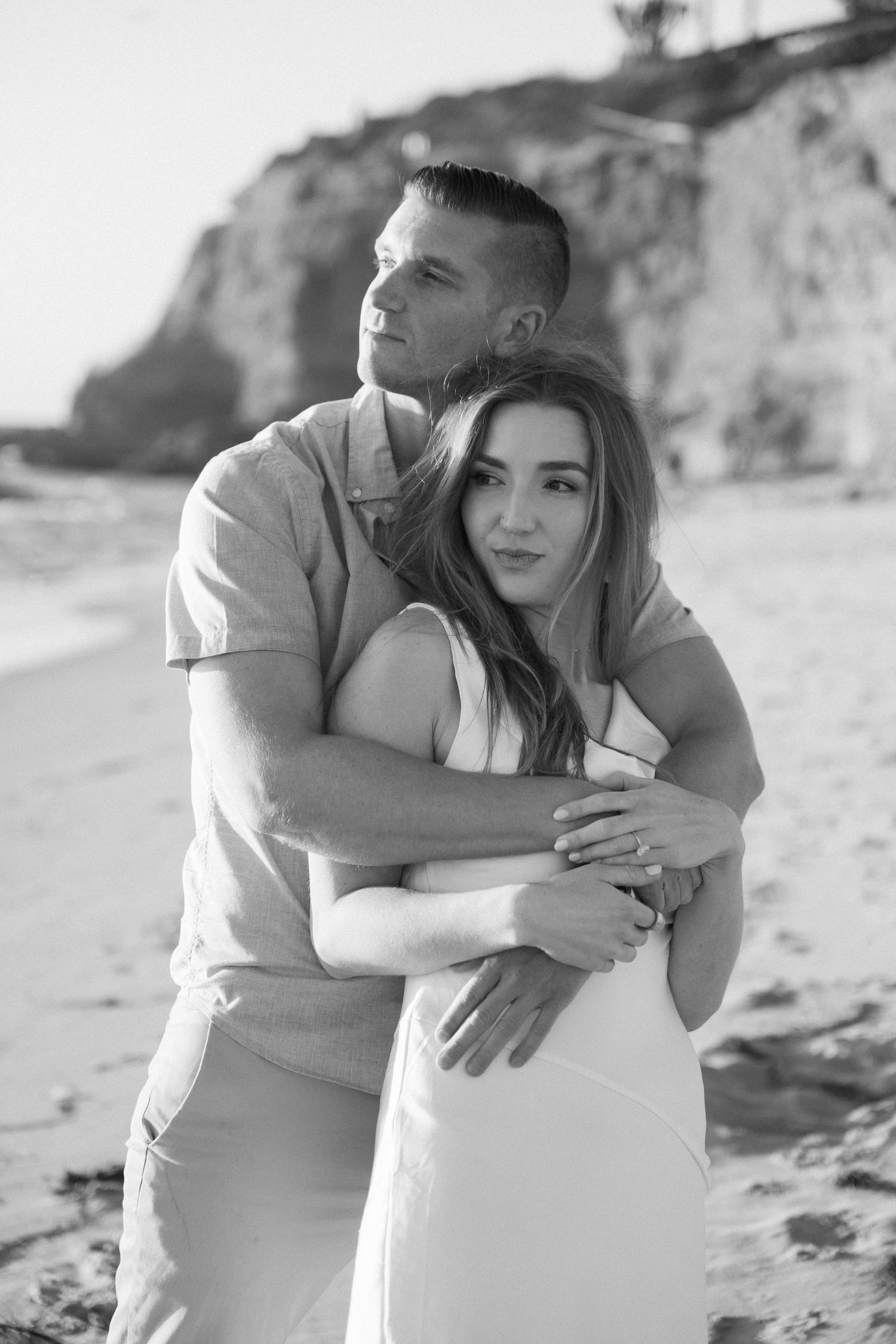 Black and white photo of a couple embracing on a sandy beach with rocky cliffs, man hugging woman from behind.