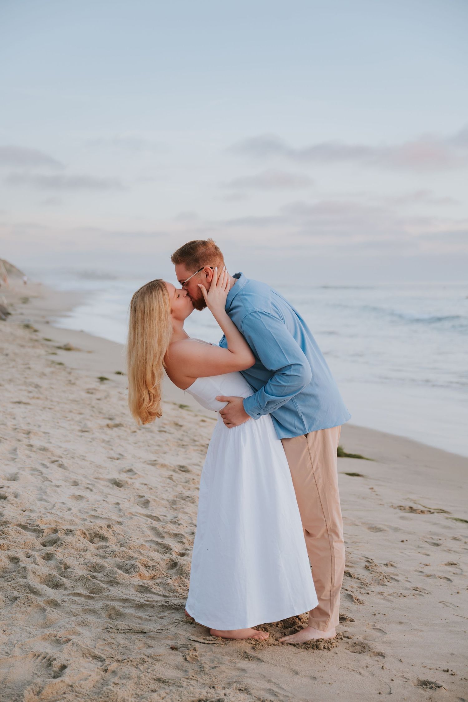 A couple shares a romantic kiss on a sandy beach at sunset, woman in white dress, man in blue shirt.