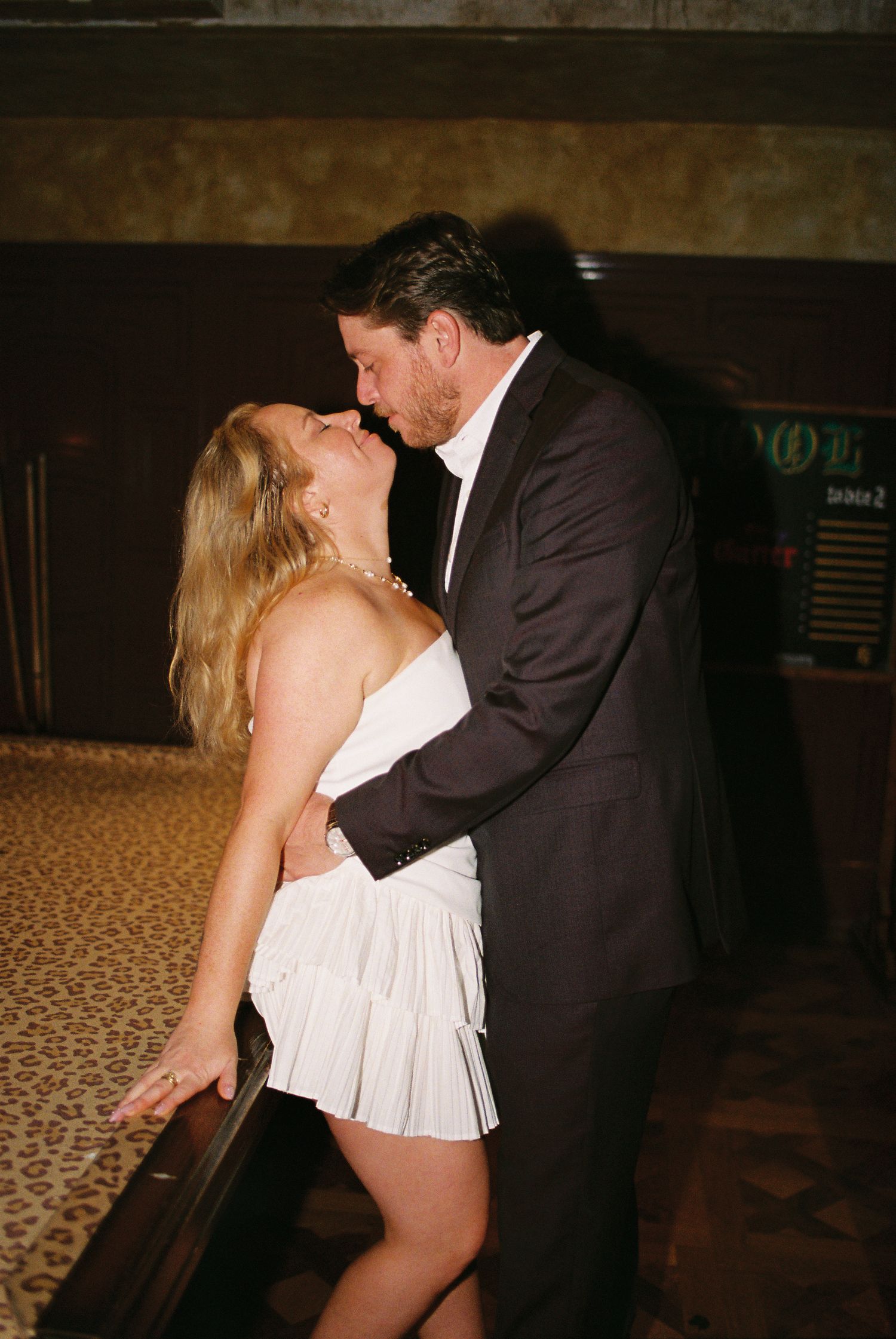 Couple sharing a passionate kiss in a dimly lit elegant venue, woman in white ruffled mini dress.