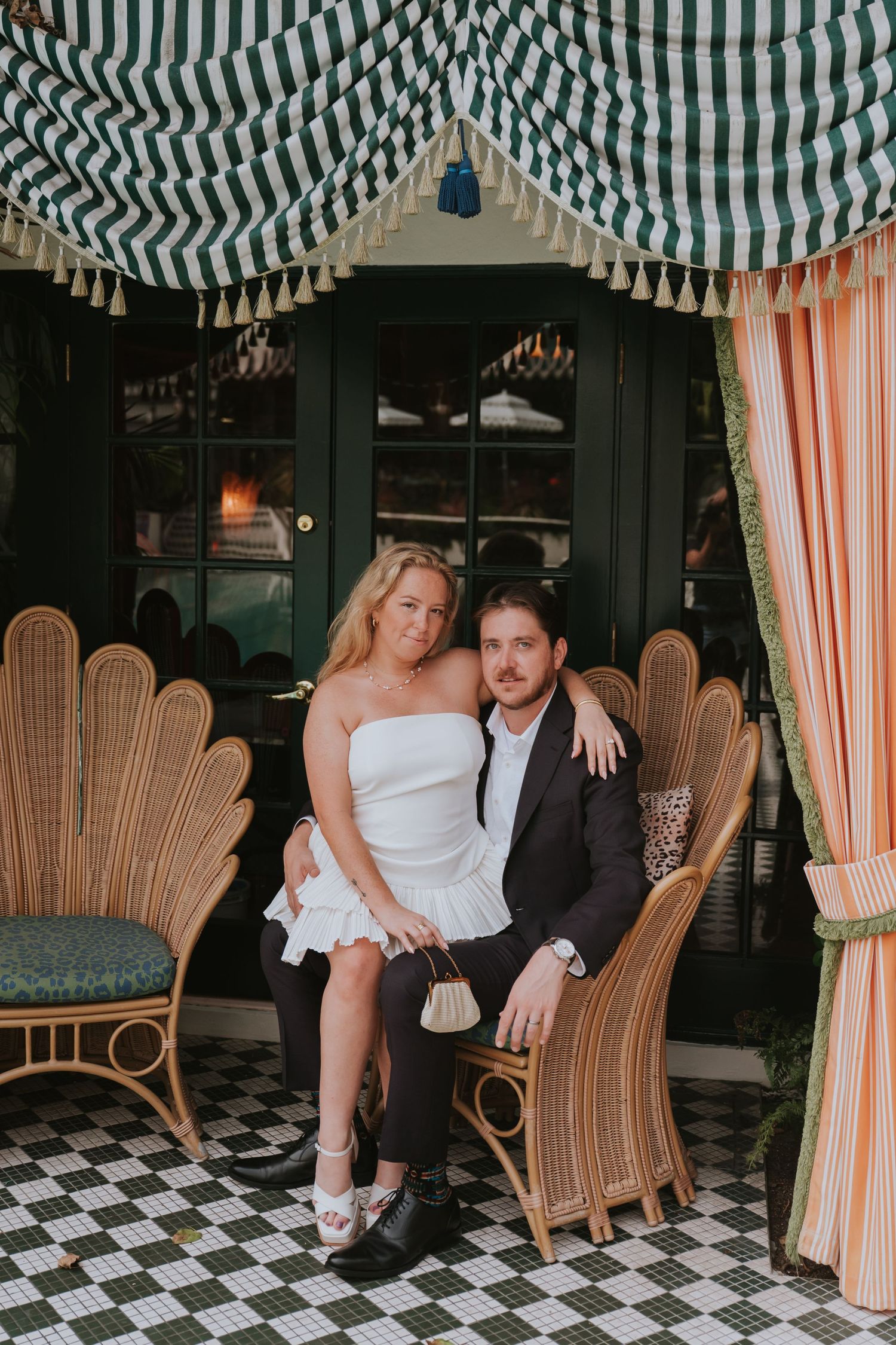 Couple posing together in elegant rattan chairs under striped green awning at stylish restaurant patio.