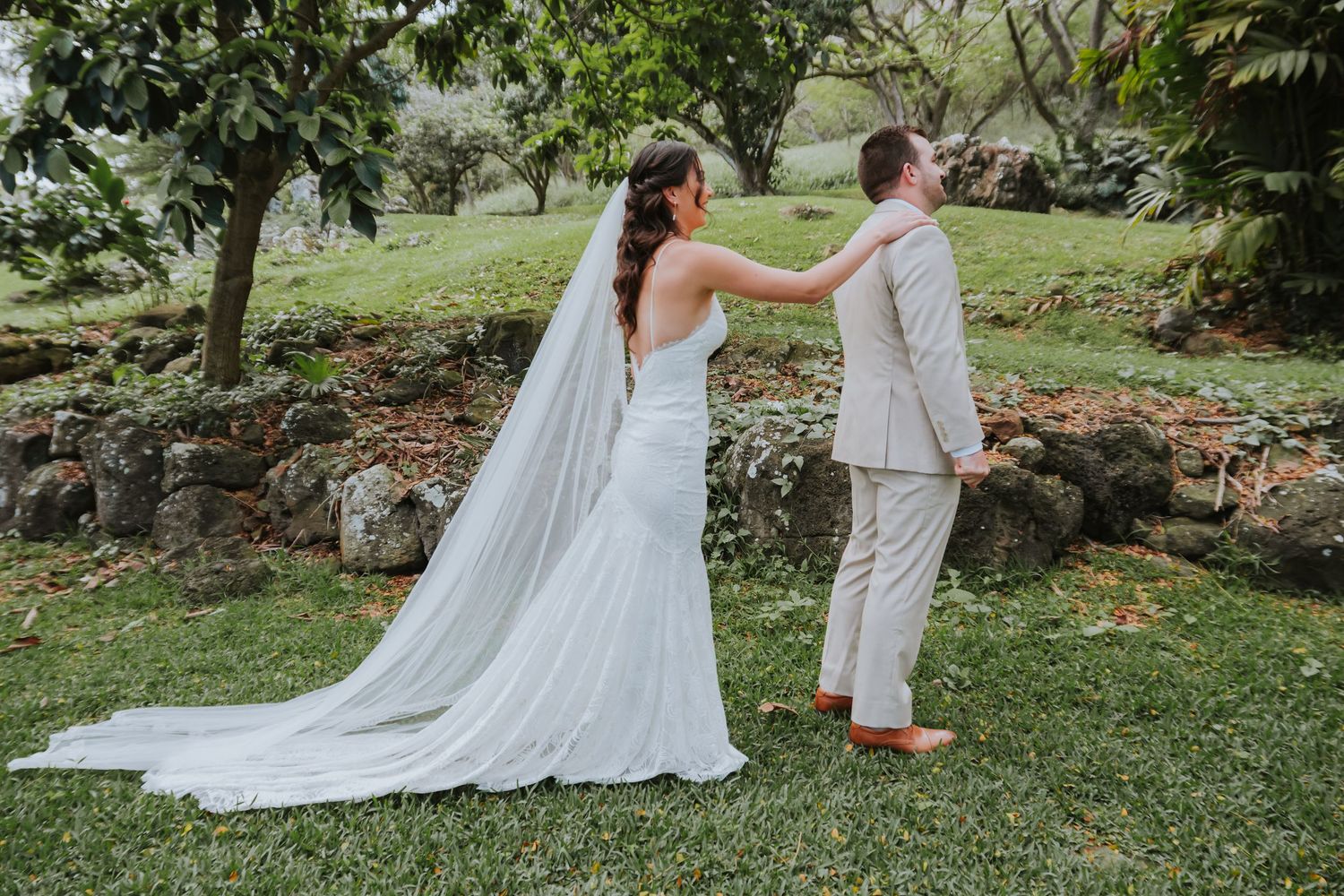 A couple in wedding attire stands together in a lush garden setting with a flowing white dress train.