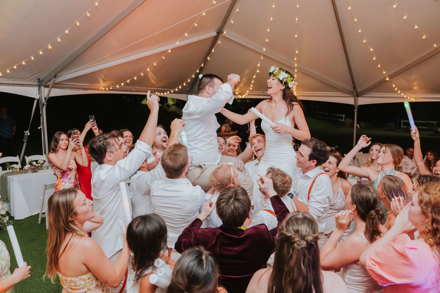 Wedding reception celebration under tent with string lights as guests lift couple in chairs.