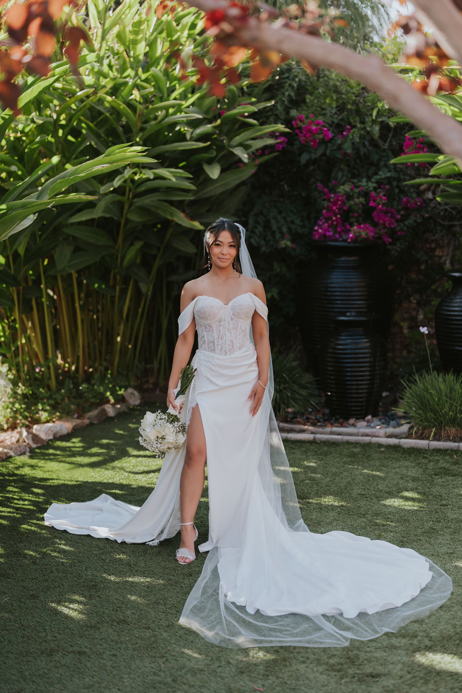A beautiful white wedding dress with a flowing train is displayed in a garden setting with lush greenery and flowers.