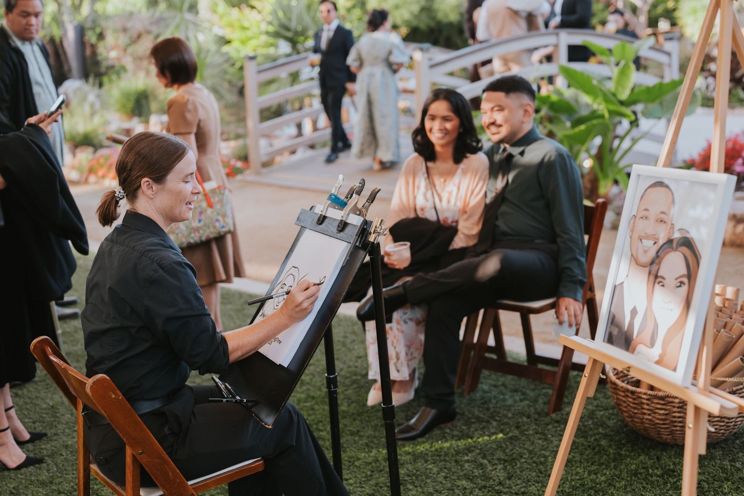Artist sketches a couple at an outdoor wedding event while displayed portraits rest on easels in the background.