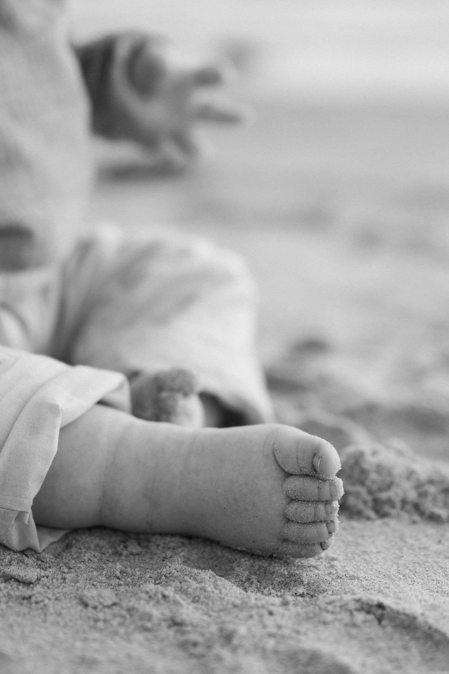 Close-up black and white photo of a baby's tiny feet resting in soft sand at the beach.