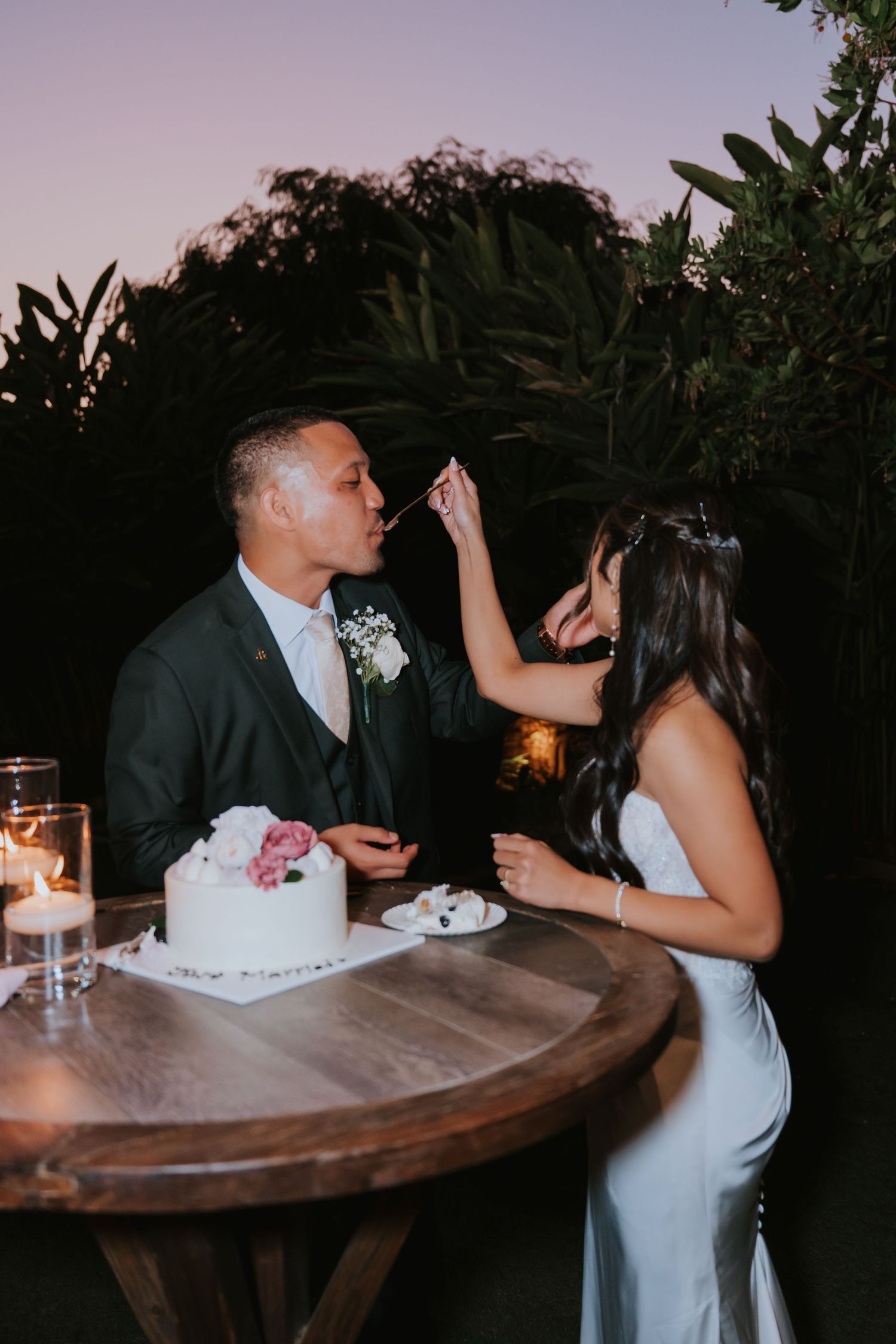 Newlyweds enjoy cake feeding moments at a rustic wooden table during sunset.