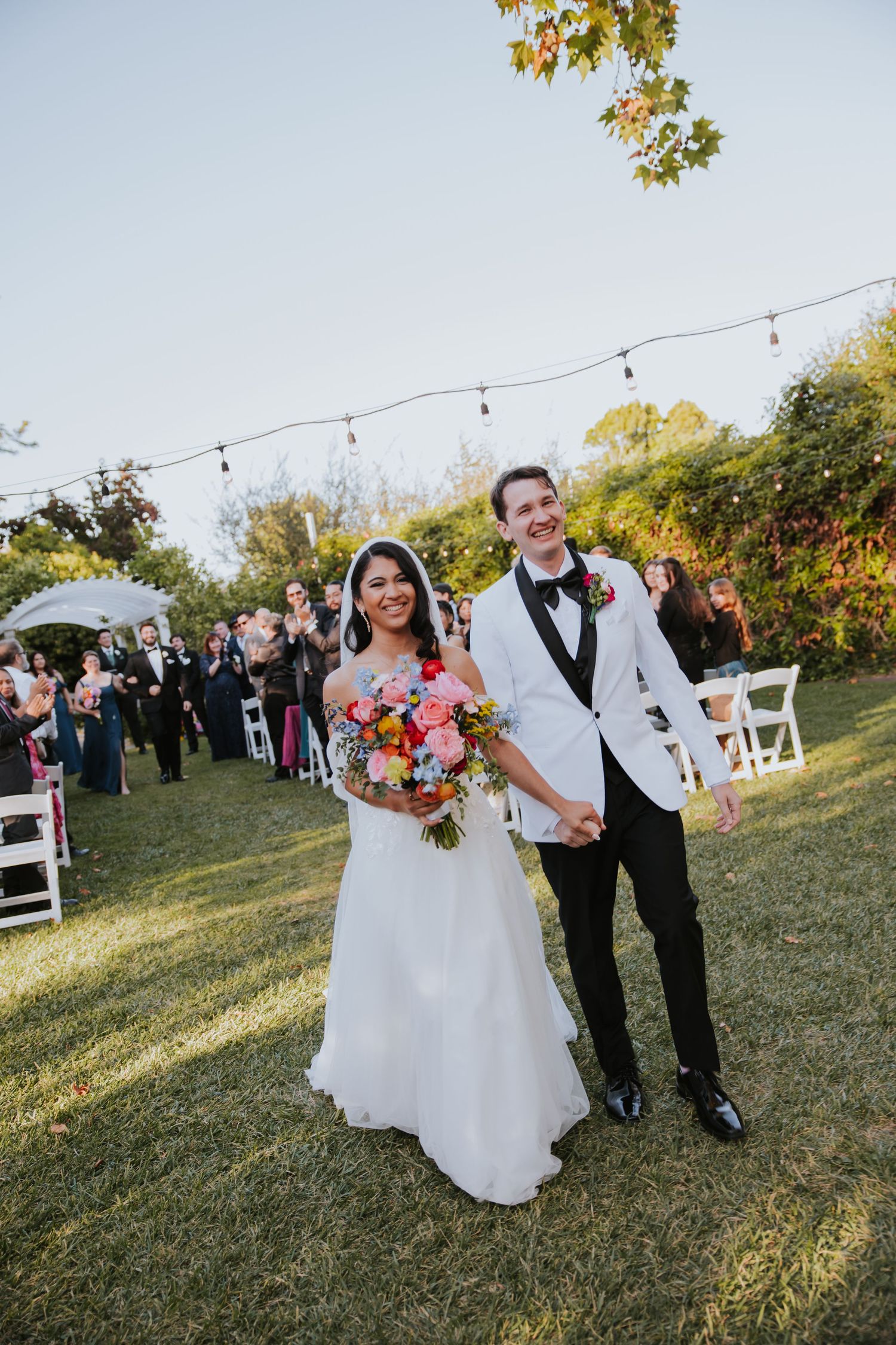 Newlyweds celebrating with their wedding party on a grassy lawn with string lights overhead.