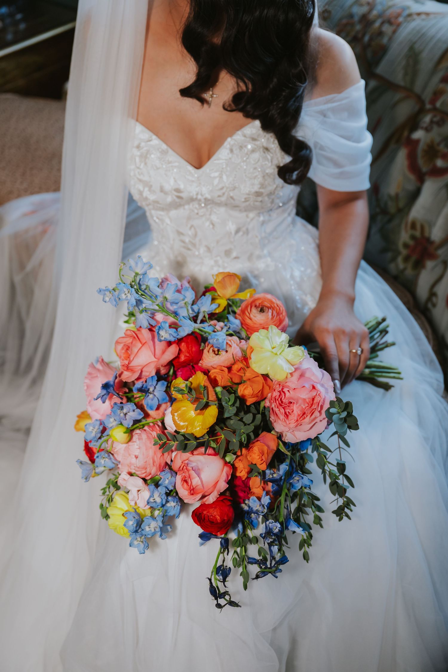 A vibrant wedding bouquet with coral peonies, blue forget-me-nots, and yellow blooms against a white bridal gown.