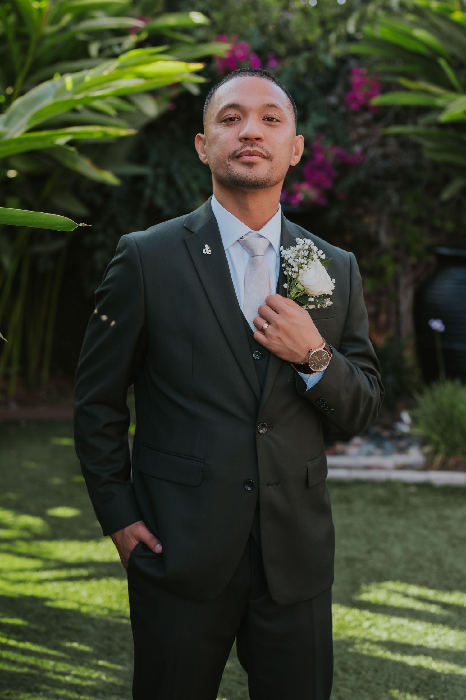 Person in a black formal suit adjusts their tie while standing in a tropical garden setting.