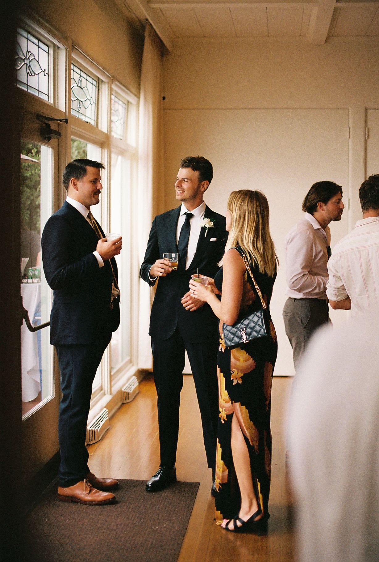 Guests in formal attire socializing indoors at a wedding reception, holding drinks near large windows.