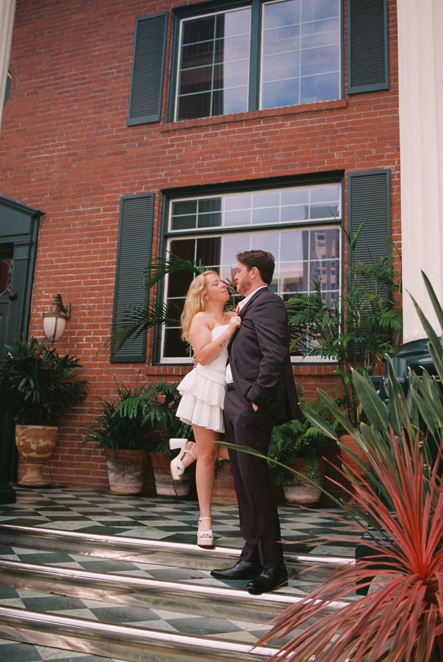 Couple in wedding attire embracing on steps of a brick building with green shutters and lush potted plants.