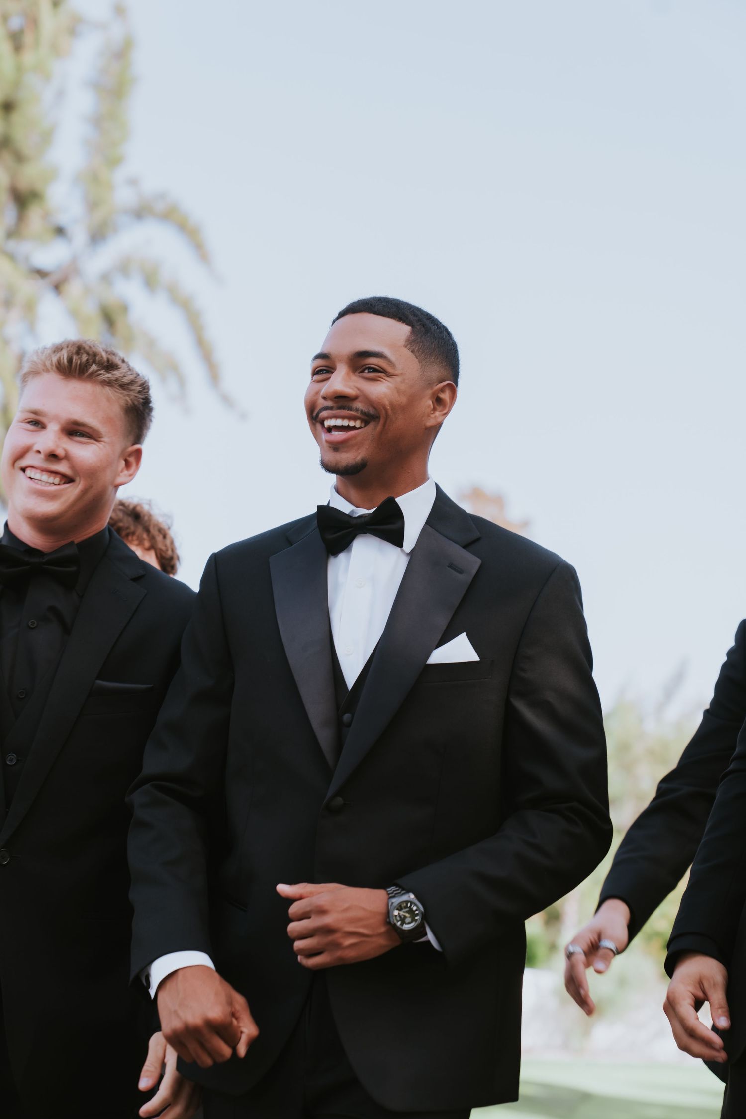 Men in black tuxedos smiling outdoors at a formal event, with palm trees in the background.