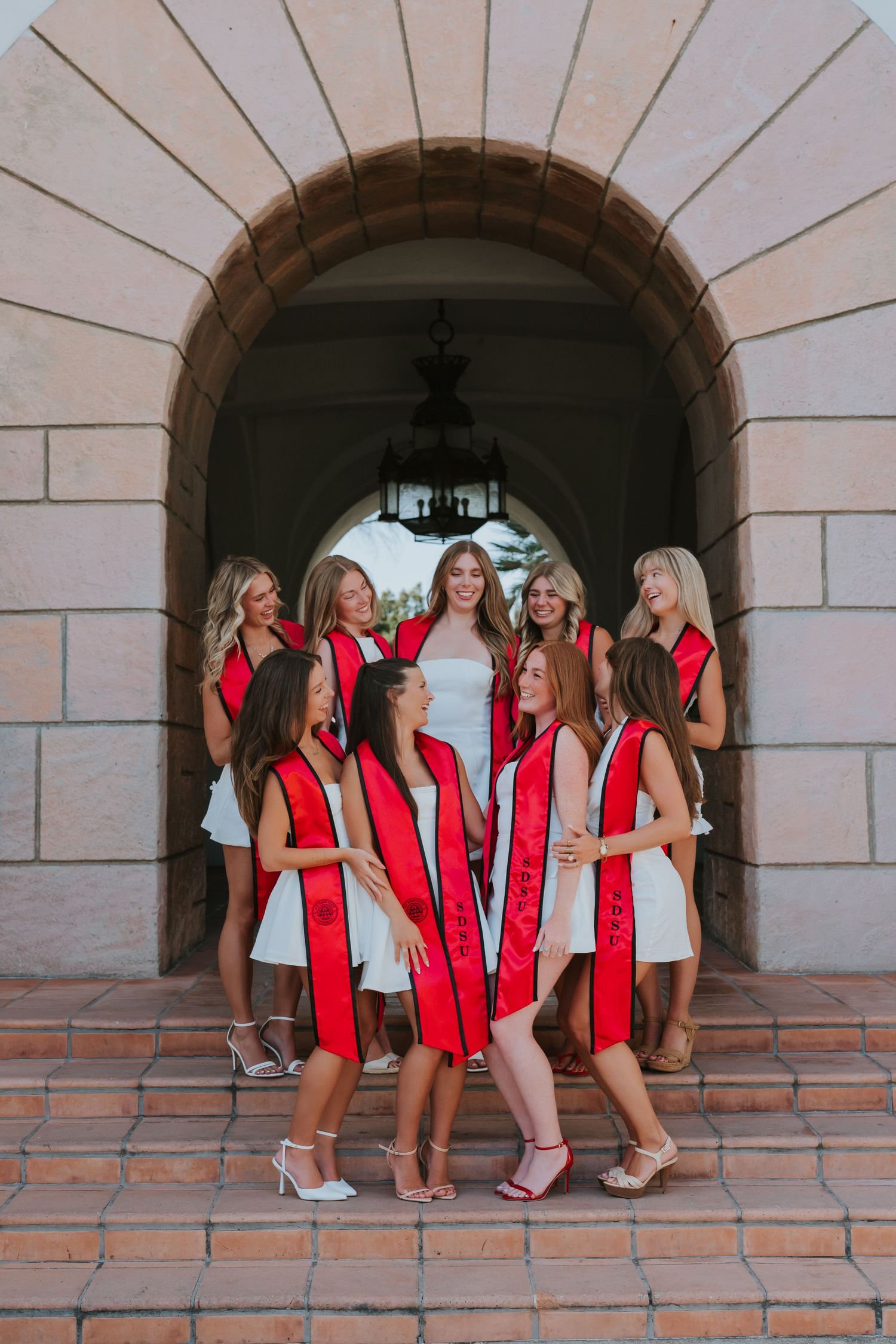 Group of young women in white dresses with red graduation sashes posing under a stone archway.