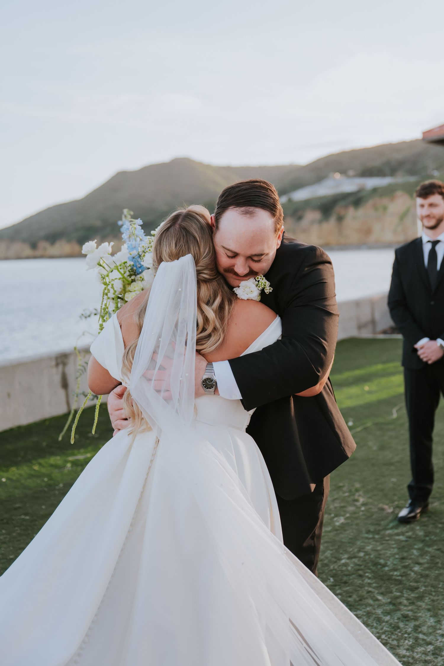 Bride and groom share an emotional hug during outdoor wedding ceremony with ocean and mountains in background.